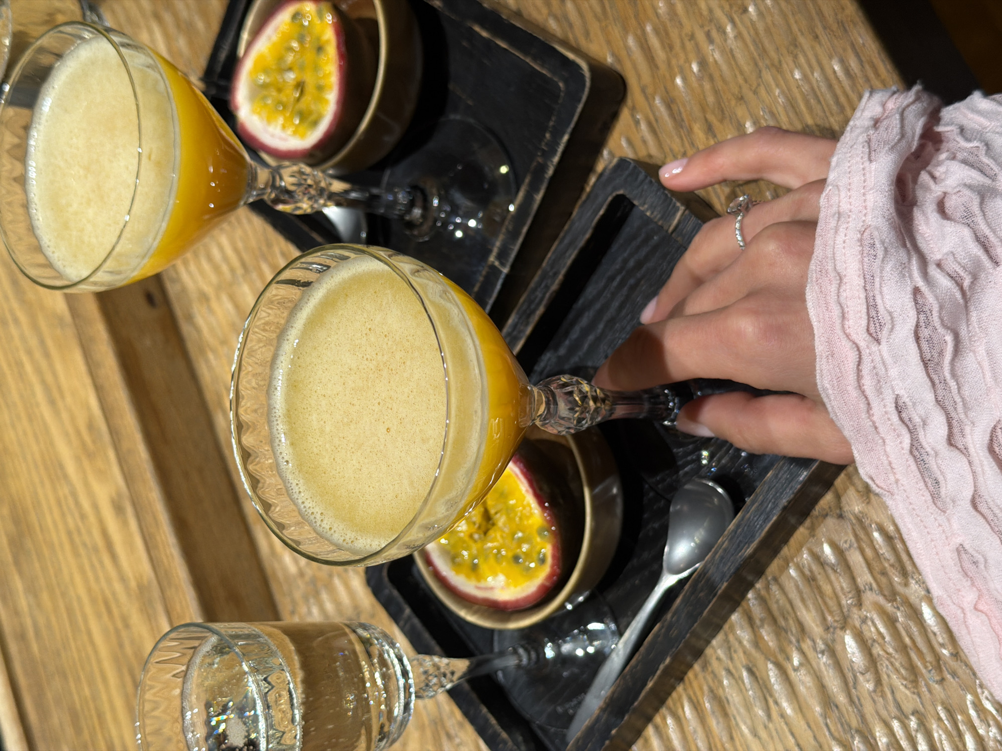 An overhead shot captures a tabletop scene featuring three beverages and a person's hands. Two stemmed glasses contain a frothy, opaque yellow-orange drink, possibly a cocktail or juice. A third stemmed glass holds a clear, sparkling beverage.
The drinks are presented on two separate dark, textured wooden trays. Each tray holds one of the yellow-orange drinks, a small bowl containing a half of a passion fruit with its vibrant yellow pulp and seeds, and a small silver spoon. The passion fruit halves suggest they are intended to be scooped out and enjoyed.
A person's hands are visible on the right side of the frame, reaching towards or holding one of the glasses with the yellow drink. The person wears a light pink, textured sweater or sleeve. A silver ring with a clear, sparkling stone is visible on one of the fingers. The nails are neatly manicured with a light polish.
The table surface is a light-colored wood with a pronounced grain and subtle texture, likely within an indoor setting such as a restaurant, cafe, or home. The overall presentation suggests a leisurely occasion, such as brunch or a social gathering. No specific time of day or weather conditions are discernible, and no text is visible within the image. The location context provided for this scene is Krasnoyarsk, Russia.