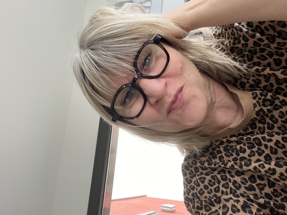 An adult female is depicted indoors in Chambœuf, France, looking directly at the camera. She has blonde hair with visible grey roots and bangs, wears black, somewhat hexagonal-framed glasses, and a brown and black leopard print t-shirt. Her left hand or arm is positioned near the left side of her head. The background on the left features a window revealing a portion of an exterior red-orange building with a white rectangular element, against a bright, possibly overcast sky. A white ceiling and wall are visible above her. A faint, partially obscured white object with dark outlines is present on the far right.