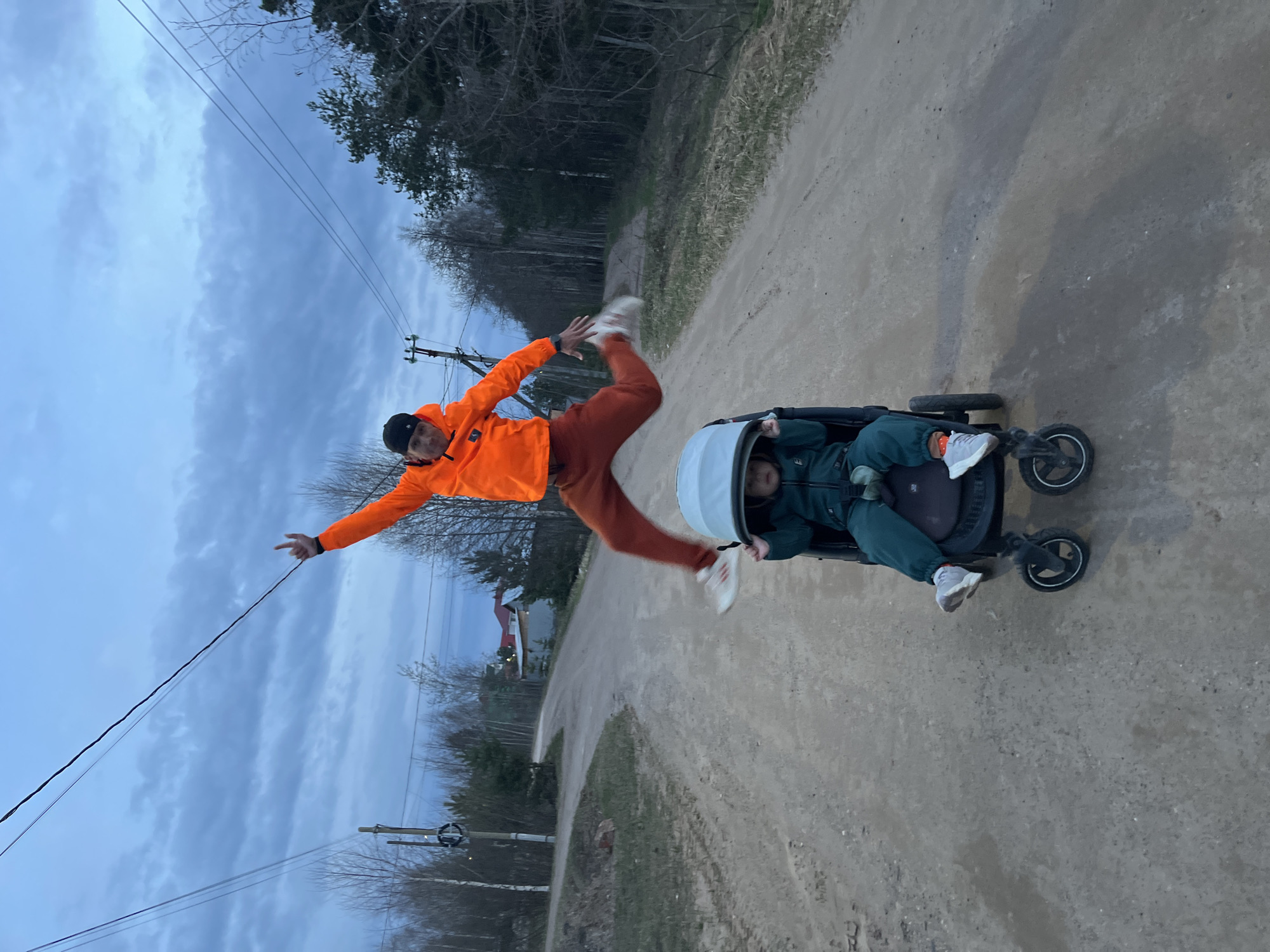 This outdoor image, taken in what appears to be a rural or semi-rural setting in Chaikovsky, Russia, captures a playful moment between a man and a child on a dirt road under an overcast sky, likely during late afternoon or early evening.
The primary subject on the left is a man dressed in a vibrant orange jacket, matching orange pants, a dark beanie, and white sneakers. He is captured mid-air, striking a dynamic pose or jump with both arms outstretched and one hand making a gesture. His movement is slightly blurred, indicating action.
To the right, a child, wearing a dark teal or green outfit, is seated in a dark-colored stroller with a light-colored canopy. The child's upper face is visible, and they appear to be looking towards the man or their surroundings.
The setting includes a wide dirt or gravel road, flanked by bare-branched trees, suggesting early spring or late autumn. A house with a red roof is visible in the distance through the trees on the left. Utility poles and power lines stretch across the cloudy sky, adding to the rural aesthetic. The ground beside the road features sparse grass and some dry vegetation. The scene conveys a casual and active outdoor experience between the two individuals. There is no discernible text visible in the image.
