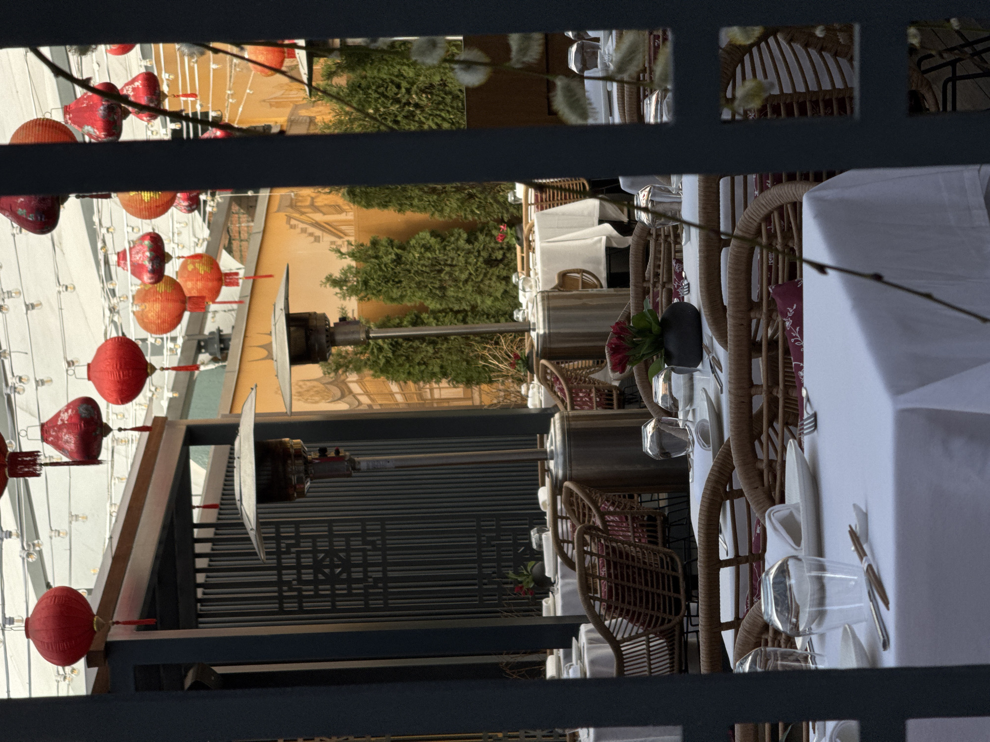 The image shows an outdoor restaurant scene in Moscow, Russia, viewed through a partially obscured, dark, square-patterned metal frame. Red Chinese lanterns are strung above the restaurant, adding a festive atmosphere. The outdoor seating area is set with tables covered in white tablecloths and surrounded by wicker chairs. Place settings include glassware and silverware. A large, dark-colored heater stands nearby, presumably to provide warmth for diners. In the background, there is a building with an ornate, painted facade and landscaping including trees.