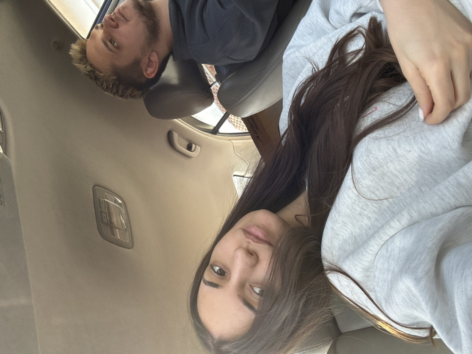 The scene depicts the interior of a passenger vehicle, identified as being in Golyamo Belovo, Bulgaria. Two individuals are present.
In the foreground, occupying the front passenger seat, is a female subject with long, dark brown hair parted on the left, cascading over her left shoulder and chest. She is wearing a light grey crew-neck sweatshirt. Her left hand, with light-colored nail polish, rests near her chest. She faces directly towards the camera, with a neutral expression.
In the background, to the right, is a male subject occupying the driver's seat. He has short, spiky, light-colored hair and visible facial stubble. He is wearing a dark-colored hooded garment. His head is turned upwards and to the right, directing his gaze outside the vehicle's frame.
The vehicle's interior features a light beige headliner with a clear plastic dome light fixture centrally located. A beige grab handle is visible above the passenger-side door. Dark-colored car seats with headrests are present. In the background, behind the passenger seat, a brown cardboard box with the partial inscription "Airtac" is discernible. Portions of the vehicle's windows reveal diffuse daylight and hints of an exterior building structure. The female subject appears to be taking a photograph. No direct interaction between the subjects is evident.