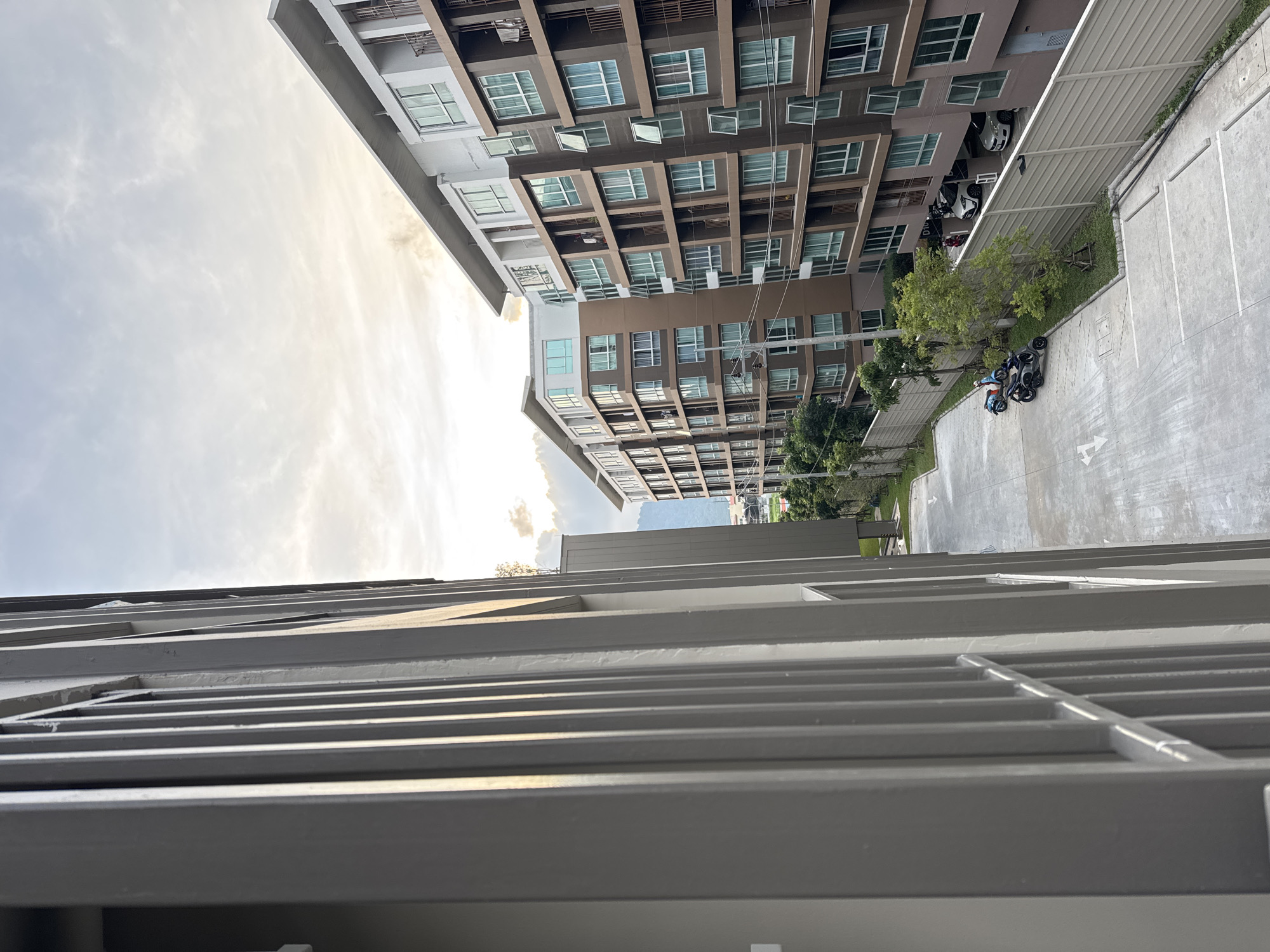 A view from a balcony looks out over a courtyard area adjacent to two apartment buildings. The buildings are multi-story structures with brown exteriors and numerous windows. In the courtyard, there are parked vehicles including two scooters, and two cars parked under the overhang of one of the buildings. A concrete parking lot with painted white lines is visible. Trees and landscaping are interspersed throughout the courtyard and along the perimeter of the buildings. The sky is cloudy, with sunlight breaking through. The scene is located in Kathu, Thailand.