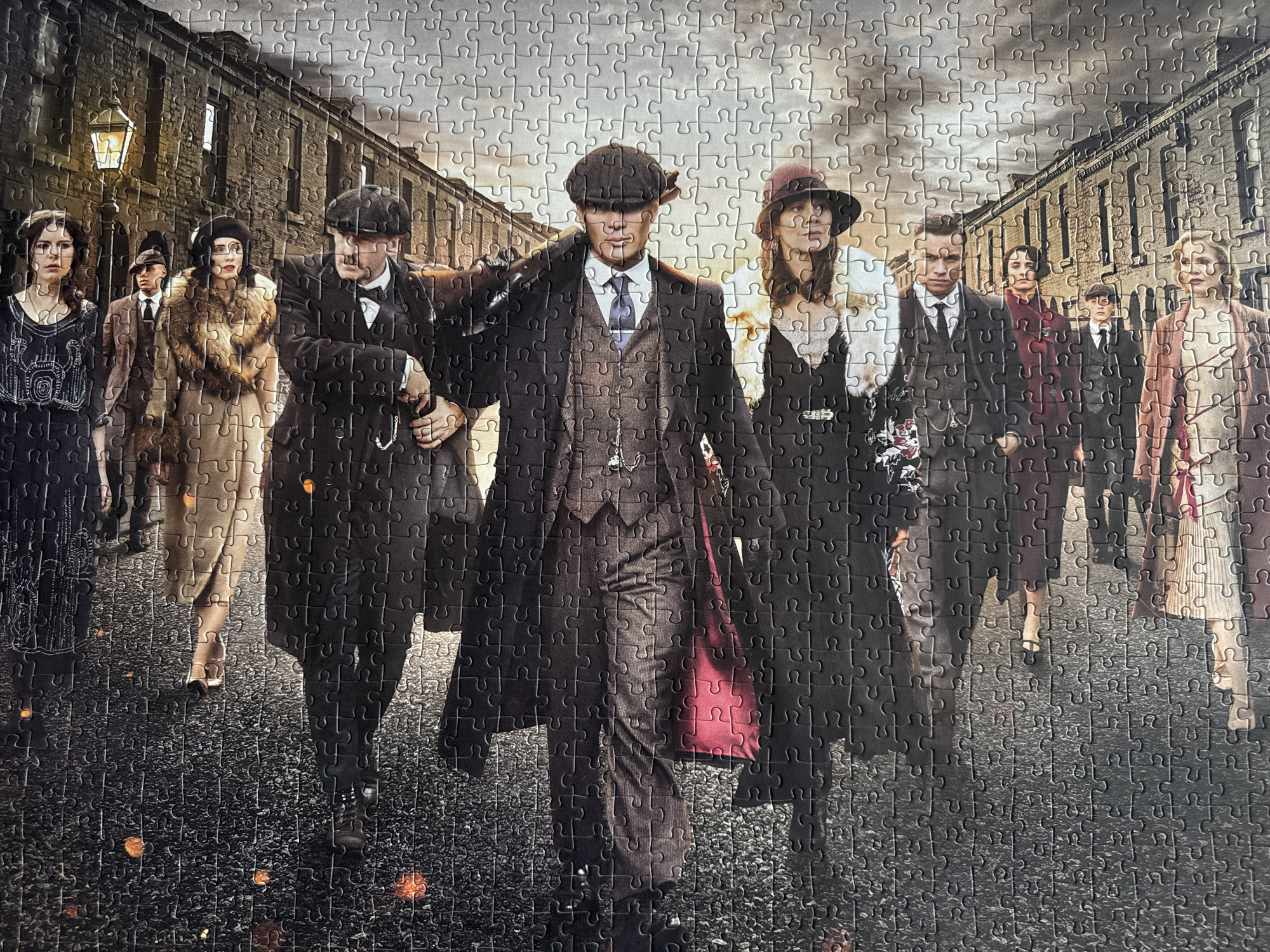 The image is a photograph of a jigsaw puzzle depicting a group of people in vintage clothing. The scene appears to be set in a city at night, with a lamppost visible in the background and some orange glowing embers on the ground. The people are arranged in a line, some looking at the camera and others looking away. They are all dressed in suits, hats, and dresses that suggest a historical period, possibly the early 20th century. One person in the foreground is holding a cigarette. The puzzle itself is laid out on a surface, with its pieces clearly visible. The image does not contain any information about the city of Craigavon, United Kingdom.
