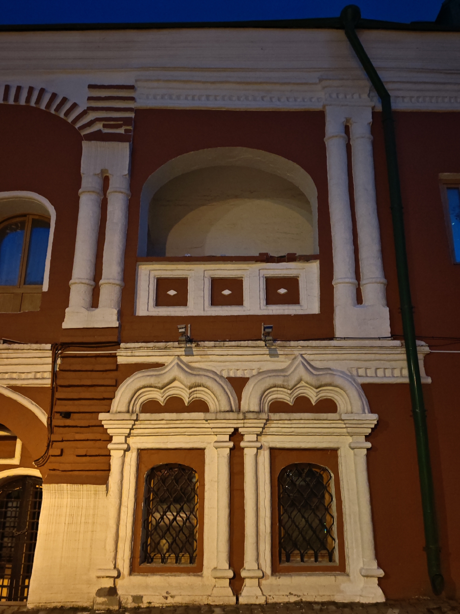 A three-story red building with white architectural details. The top floor has a row of arched openings with white trim, columns, and details. The second floor features a niche or balcony-like structure with three decorative diamond-shaped insets below the arched opening. The first floor displays two arched windows with white trim and decorative elements above each window. A green drainpipe runs vertically along the wall. The building is illuminated from below, casting shadows and highlighting the architectural details. The sky above is dark blue. The building is located in Moscow, Russia, specifically in the historical center near Red Square.