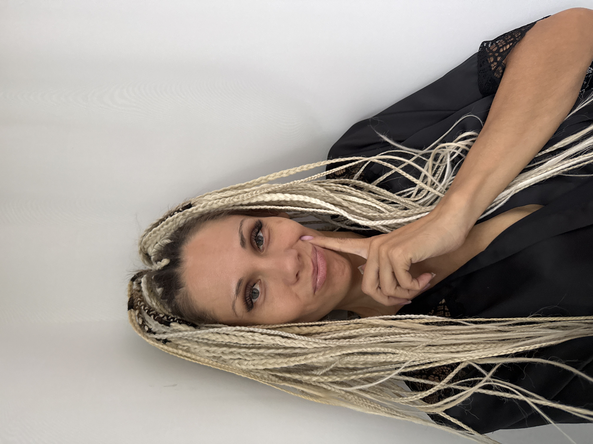 The image shows a woman with blonde braids in Yuryatino, Russia. She is wearing a black robe with lace trim and has her finger on her chin, looking at the camera with a slightly pursed mouth. The background is a plain white wall.