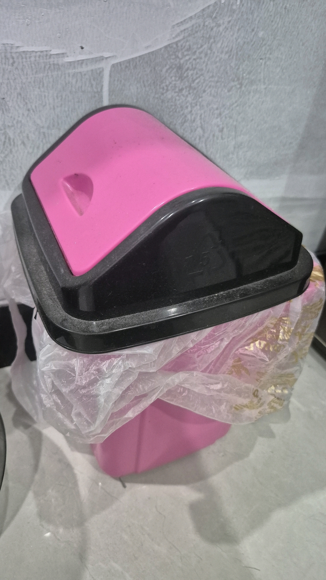 The image shows a close-up view of a pink and black trash can with a clear plastic bag partially covering the top opening. The trash can has a curved, pink lid with a black rim and a black, slanted flap. The plastic bag appears to be tied at the top and hangs over the sides of the trash can. The background consists of a textured, light-colored wall, possibly painted or wallpapered, with some faint lines or marks visible. The surface beneath the trash can appears to be a light-colored, possibly tiled floor. No people or actions are depicted in the image. The scene appears to be indoors, possibly in a domestic or public space. There is no indication of the location being in Gambiranom, Indonesia, as the image itself provides no such contextual information.