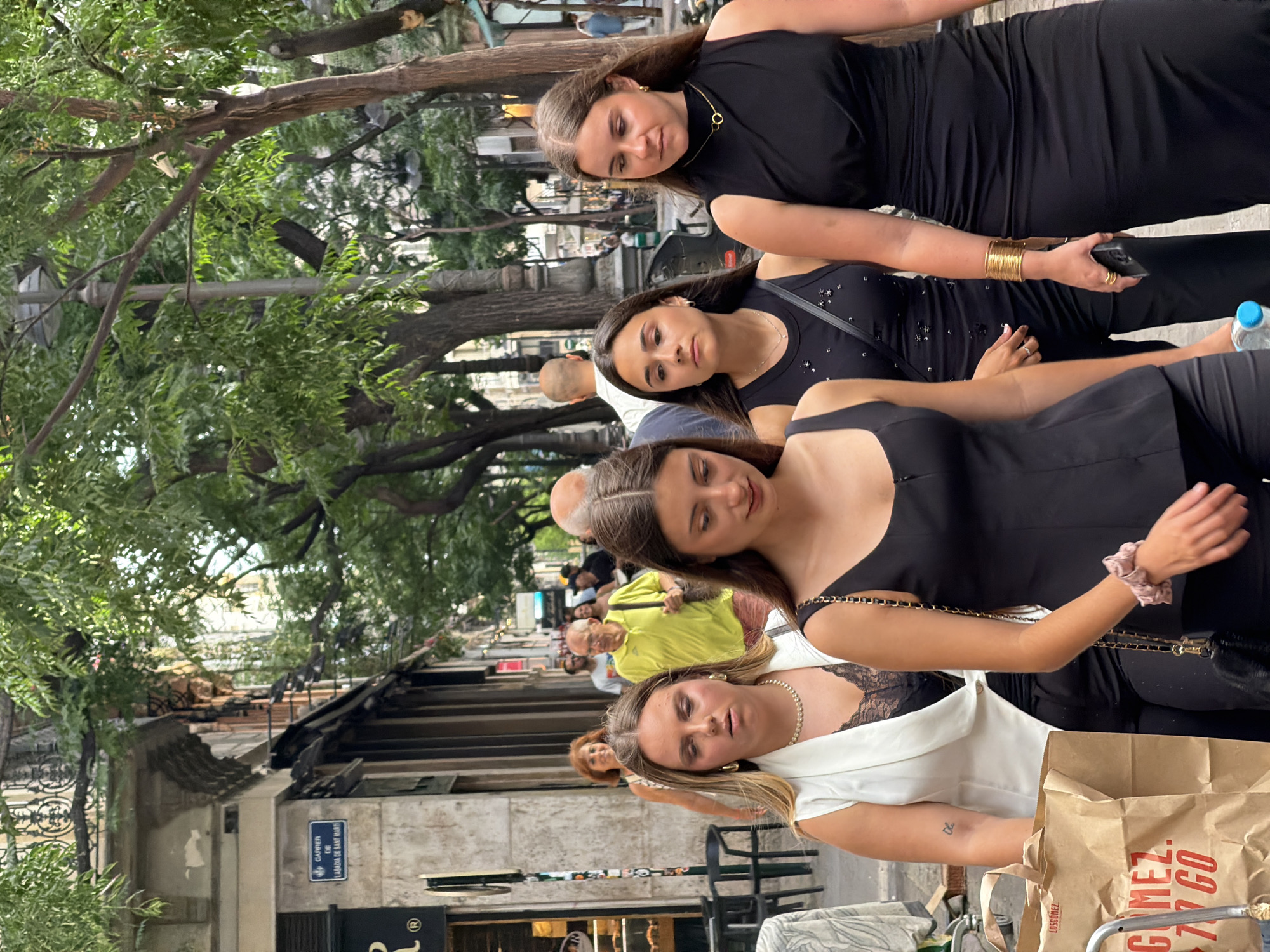 A group of people walk down a street in an urban environment. The street is lined with buildings and trees. In the foreground, four women are walking in a single file line, facing forward. The woman on the left is wearing a white vest over a black lace top and black pants. She is holding a paper bag. The second woman from the left is wearing a black sleeveless top and black pants, with a beaded necklace. The third woman from the left is wearing a black sleeveless top and black pants, with a gold bracelet on her left wrist and a ring on her right ring finger. She is holding a black smartphone in her right hand and a light blue water bottle in her left hand. The woman on the far right is wearing a black sleeveless dress and a gold necklace. In the background, several other people are walking in the same direction. A street sign is visible on the left side of the image. The street appears to be paved with stone.