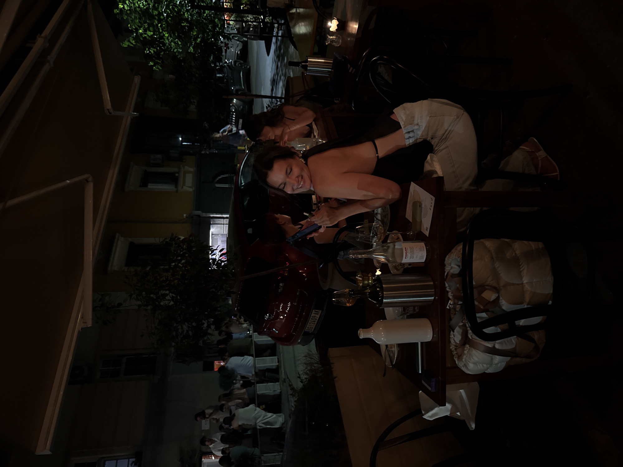 The image depicts an outdoor dining scene at night, likely in Belgrade, Serbia, given the license plate visible on a red car in the background. A group of people are seated at tables under an awning. In the foreground, a young woman with dark hair is smiling and looking over her shoulder at the camera, holding a phone. She is wearing a black top and light-colored pants. On the table in front of her are wine bottles, glasses, and other items. In the background, other patrons are visible dining and socializing, with a red car parked behind them. The environment is dimly lit, characteristic of an evening restaurant setting.