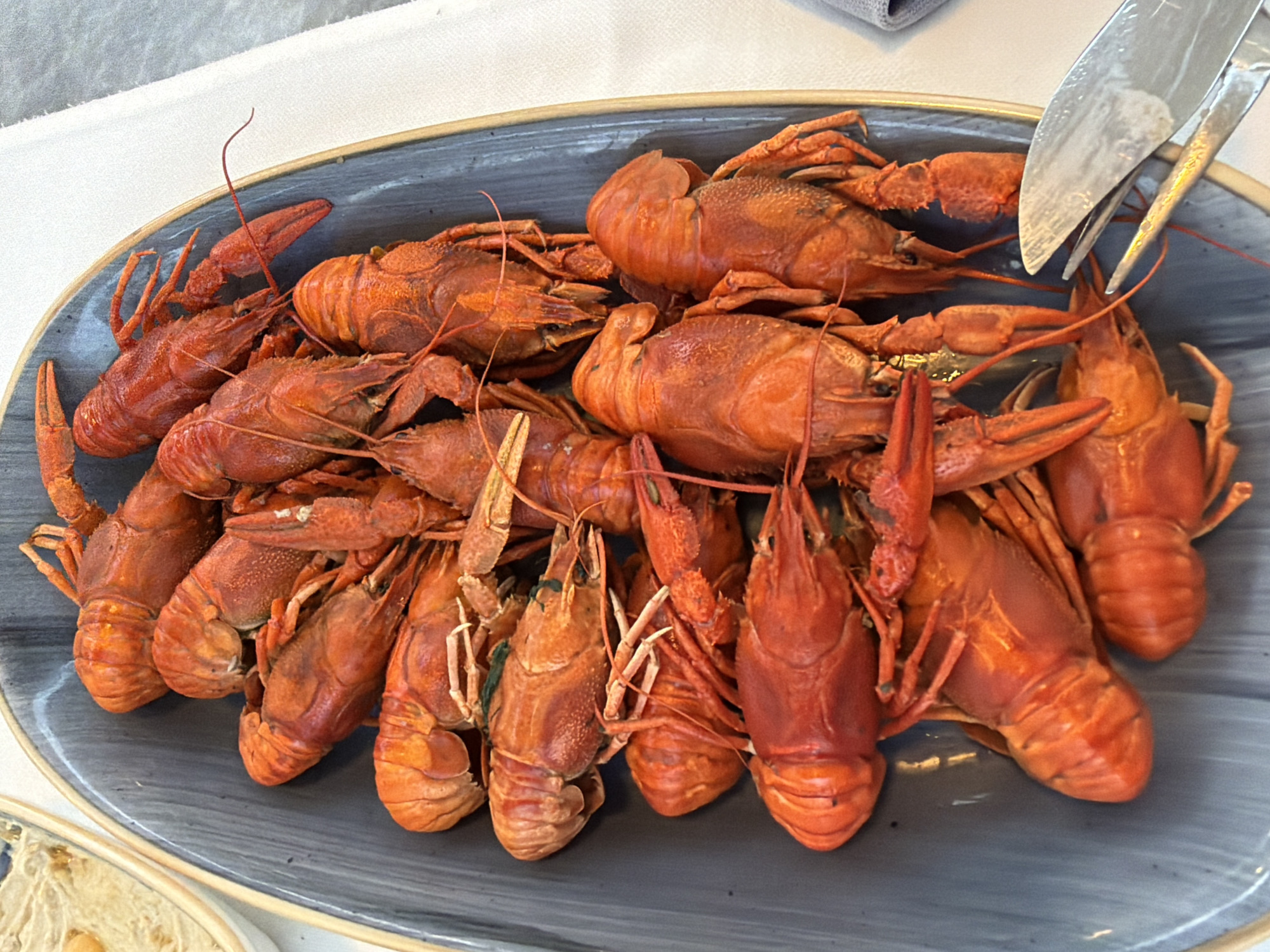 A platter of cooked crawfish sits on a table, with a white tablecloth visible to the right. The crawfish are bright red and arranged in overlapping layers. A fork and knife are positioned on the right side of the platter, suggesting the crawfish are prepared for consumption. The platter itself is dark gray and oval-shaped. To the left of the platter, a portion of another dish, possibly a dip or salad, is visible in a light-colored bowl. The overall scene appears to be a casual dining setting.