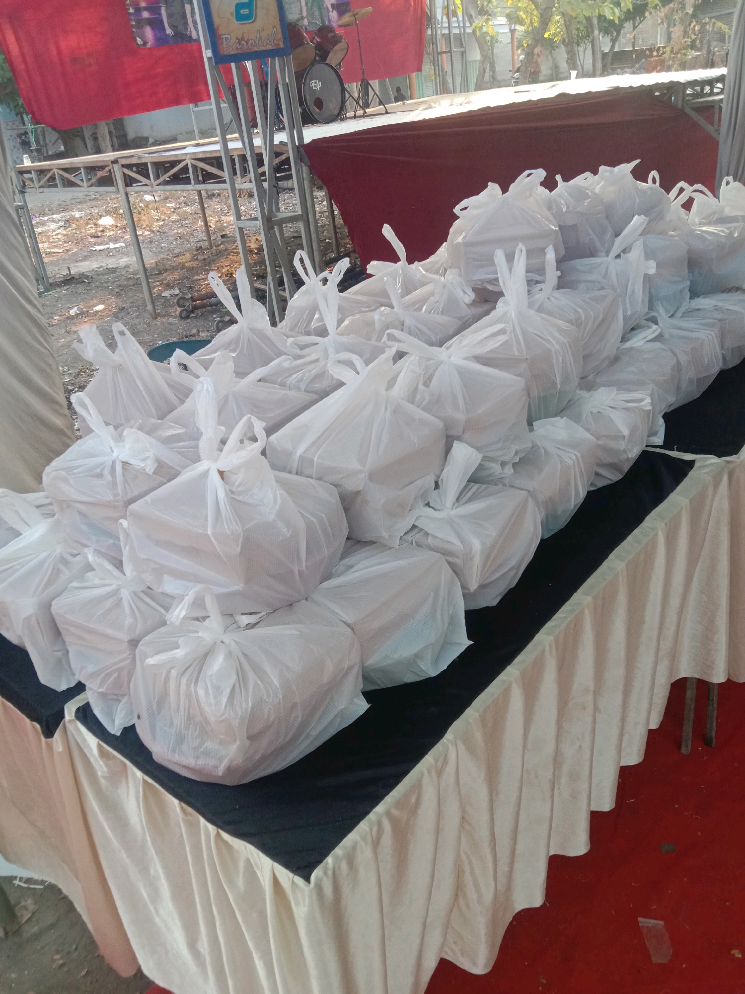 The image shows a table covered with a cream-colored tablecloth and a black cloth overlay, on which are arranged numerous white plastic bags containing food. The bags are tied at the top and vary in size and shape, suggesting they hold individual meal portions. Behind the table, a red banner hangs above a metal stage structure. A drum set is visible on the stage. The ground appears to be dirt and gravel. The overall scene suggests an outdoor event or distribution of food in Karang Anyar, Indonesia.