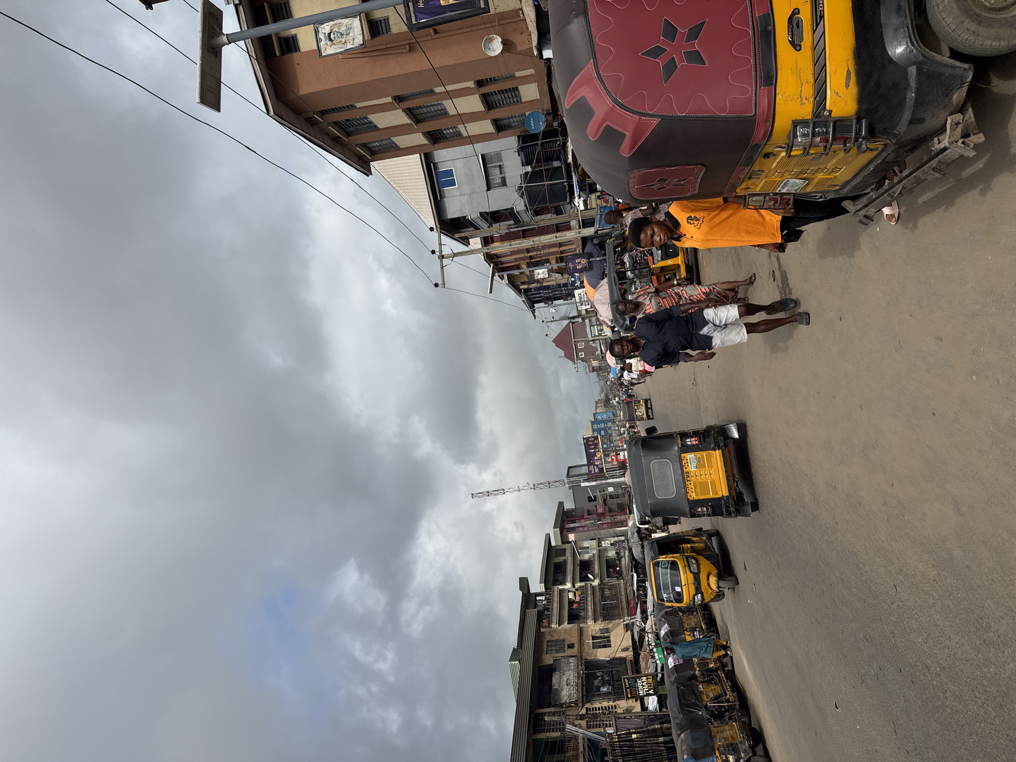 The image depicts a street scene in Fegge, Nigeria. The sky is overcast. Multiple three-wheeled vehicles, predominantly yellow, are visible on the street, some stationary and some in motion. People are present on the side of the street, some standing near the vehicles. Buildings line both sides of the street, showing signs of age and wear. Overhead, power lines and poles are visible. In the distance, a communication tower rises above the buildings.