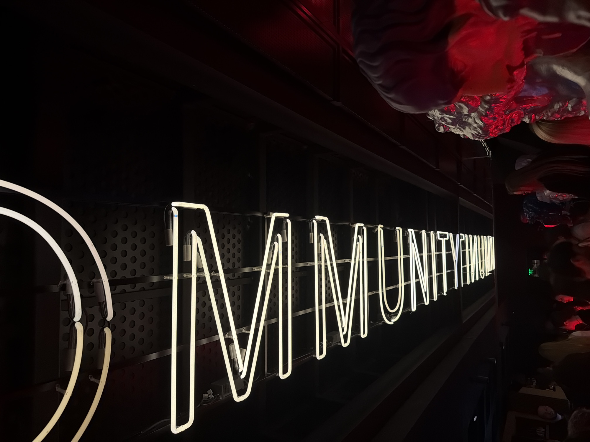 The image shows a neon sign reading "COMMUNITY MUSEUM" in Moscow, Russia. The sign is mounted on a black panel with a perforated pattern. The lighting is dim, with red and white elements in the background. There are people partially visible on the right side of the image.