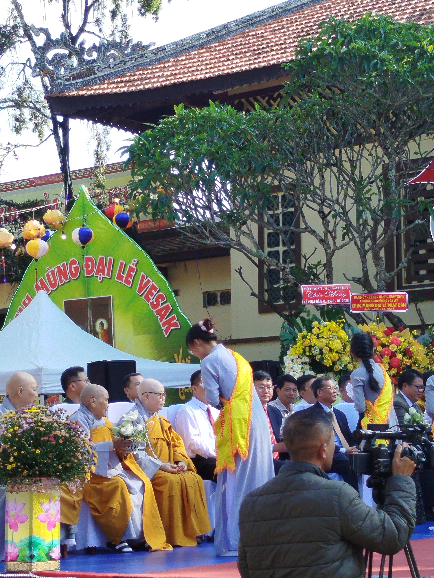 The image depicts a Buddhist ceremony in Da Lat, Vietnam. The event, labeled "Mung Dai Le Vesak," celebrating the Vesak festival, features monks in saffron robes seated on a platform. Several other people are in attendance. Behind the platform, a large green banner displays the festival's name. The background includes a building with a tiled roof. Signage displays the names of local organizations, with floral arrangements nearby. A videographer is recording the event.