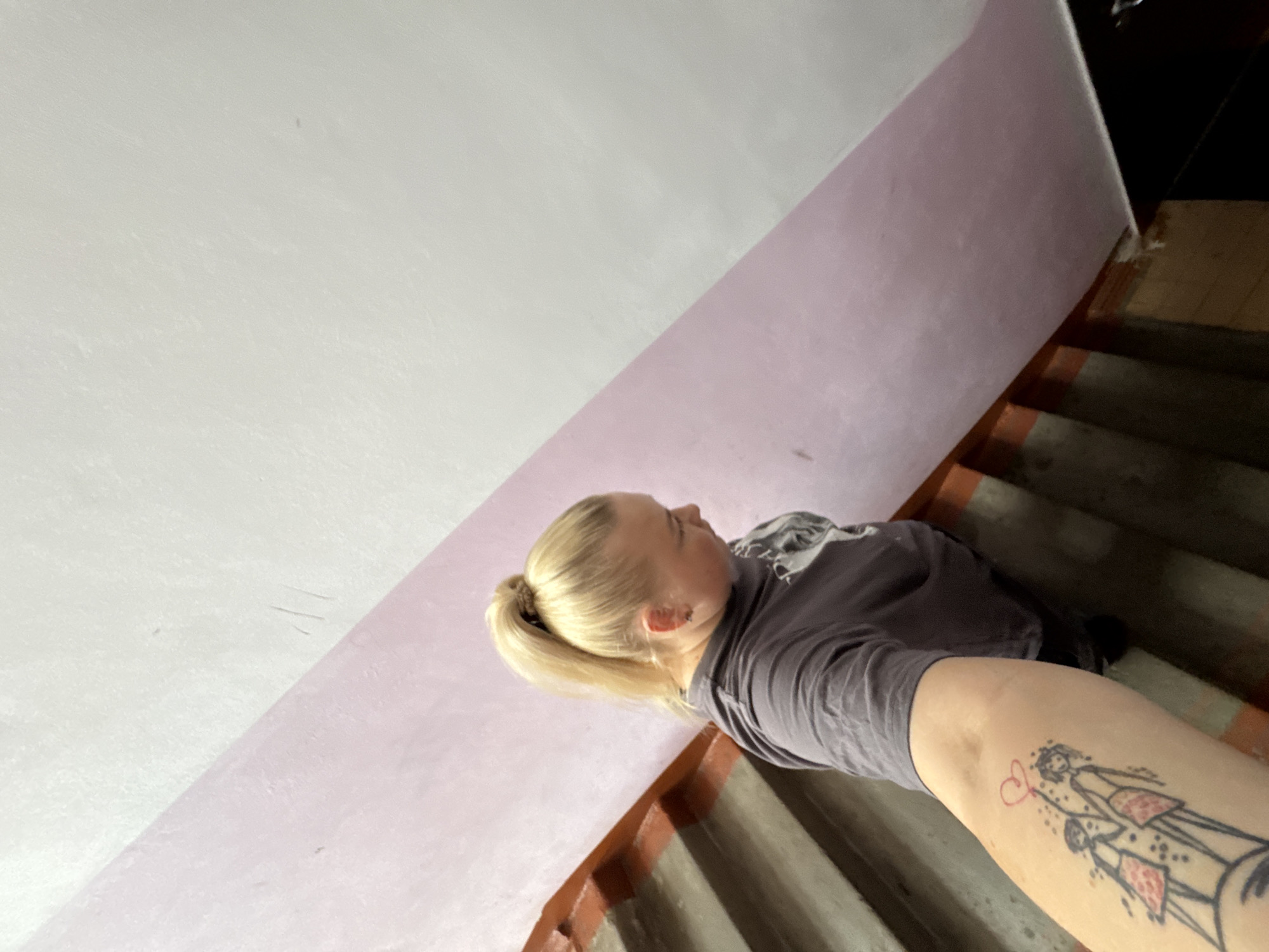A woman with blonde hair in a ponytail stands on a concrete staircase. She is wearing a grey t-shirt. A tattoo of two figures holding a heart-shaped balloon is visible on her left arm. The staircase has red painted edges. The wall next to the staircase is painted pink and white. The scene appears to be an indoor staircase, possibly in an apartment building in Chaikovsky, Russia.