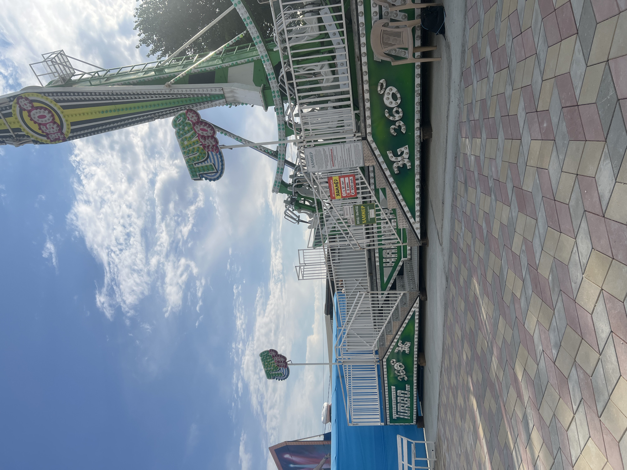 An amusement ride with a green and white structure stands on a paved area in Golubitskaya, Russia. The ride features multiple arms with seats, and one prominent arm is angled upwards towards a bright blue sky with scattered white clouds. Signs are attached to the ride structure, including one that reads "HAMMER" in yellow and red lettering. The ground is a colorful patterned pavement with a geometric design. In the foreground, two beige chairs are placed near the ride. A white bench is visible to the left of the ride, near a blue tent-like structure. The overall scene depicts an outdoor amusement park setting on a clear day.