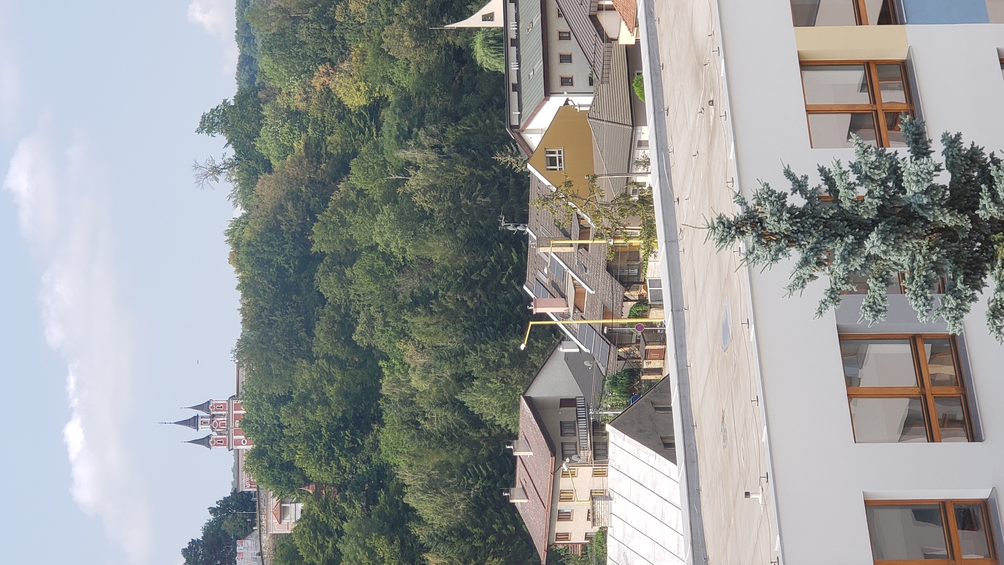 The image captures a scene in Prešov, Slovakia, featuring a residential area with houses and buildings nestled against a densely forested hill. In the background, atop the hill, a distinctive red and white building with multiple spires, likely a church or monastery, is visible. The foreground is dominated by the rooftops of several buildings, including a white-clad structure with large windows and a prominent evergreen tree. The sky above is partly cloudy. The houses in the middle ground display typical residential architecture with tiled roofs, some equipped with solar panels, and visible chimneys. A yellow street lamp stands out among the houses. The overall impression is of a quiet, semi-urban environment with a strong presence of nature.