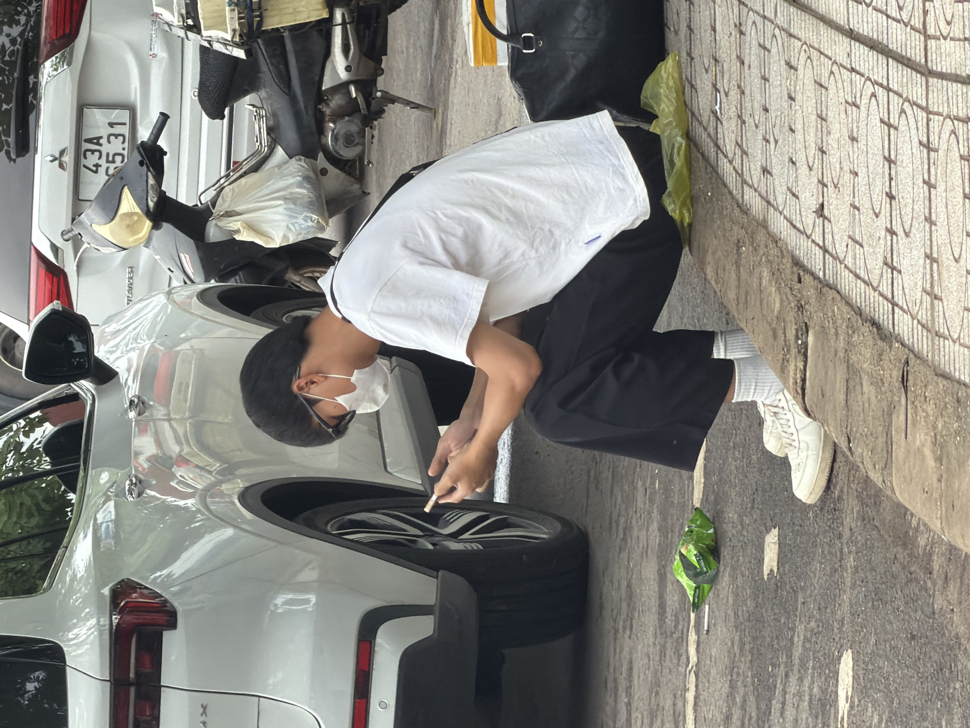 A young male wearing a white t-shirt, black pants, and white sneakers is sitting on a curb, looking at his phone. He has a white mask on his face. Next to him is a black duffel bag and a yellow plastic bag. In front of him, on the ground, is a crumpled green snack bag and a cigarette butt. Behind him, a white SUV is parked. A motorcycle with a license plate reading "43A 65.31" is also parked nearby. The scene appears to be on a street in Lao Bảo Commune, Vietnam, indicated by the presence of the motorcycle's license plate which is a Vietnam plate format. The ground is paved with concrete and tiles.