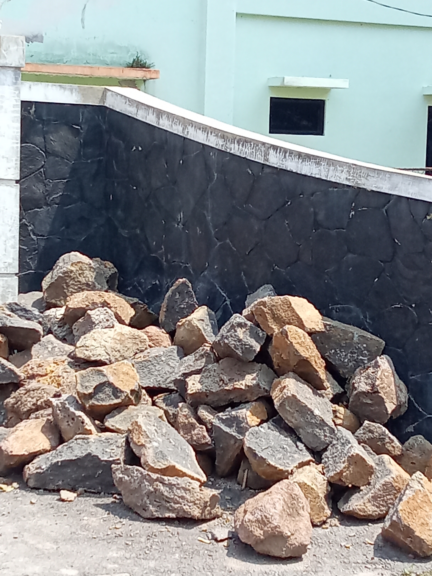 In Subang, Indonesia, a pile of rough-cut stones lies on a dusty, gray surface in front of a dark, textured wall. The wall is constructed from irregularly shaped black stones and appears to be part of a building's facade. Above the wall, a pale green building with a small, dark-paned window is visible. The stones in the foreground vary in color from light brown to dark gray, with some showing a rough, weathered texture. The scene suggests a construction or landscaping area.