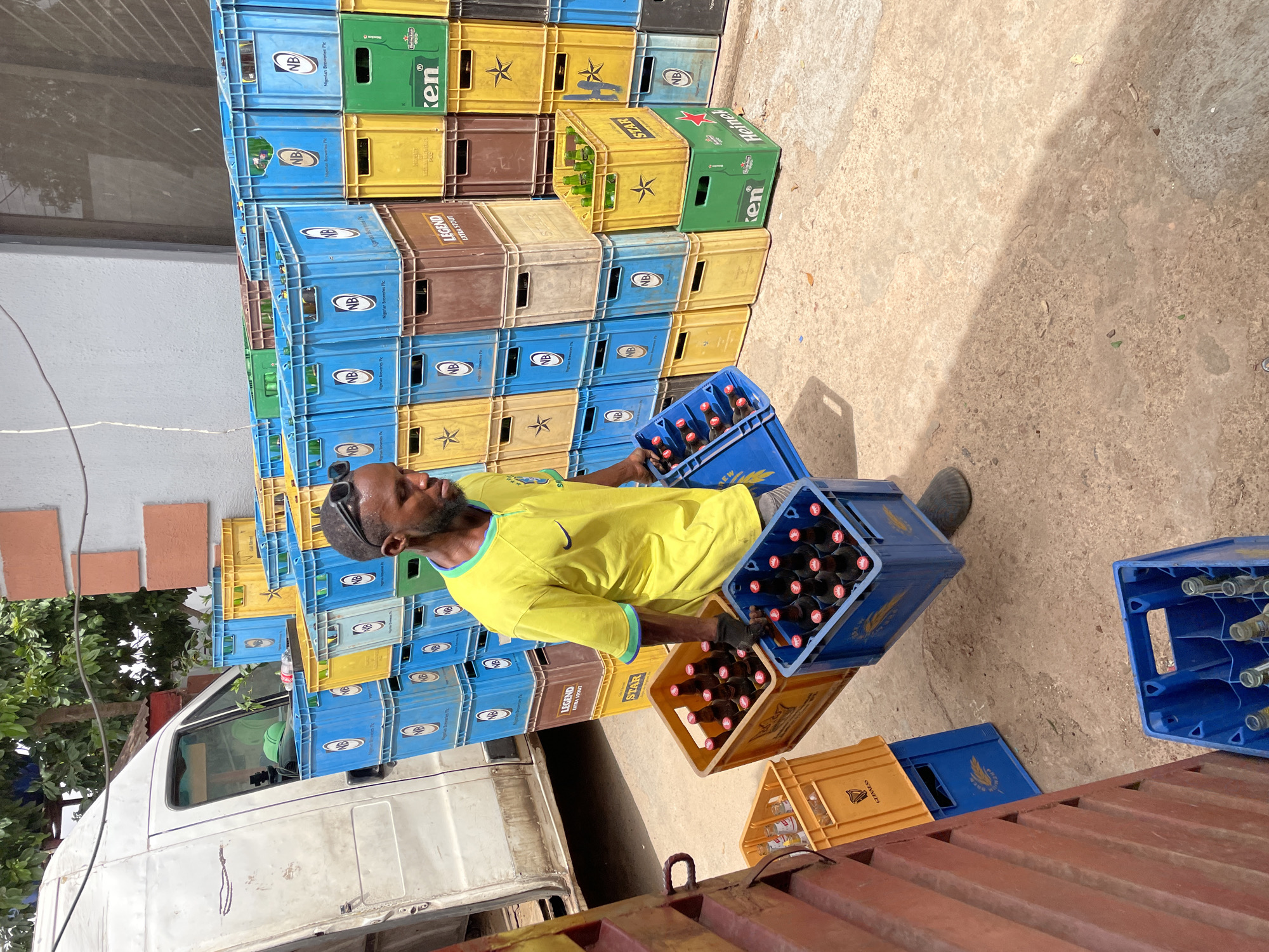 The image depicts an outdoor scene in Jos, Nigeria, focusing on a man handling beverage crates under bright daylight. An adult male, with dark skin, a short beard, and sunglasses pushed onto his head, is the central figure. He is dressed in a yellow t-shirt with light blue trim and a Nike swoosh, and wears dark gloves. The man is actively engaged in manual labor, carrying two stacked crates filled with dark bottles, likely beer or stout, suggesting he is loading or unloading inventory.
The setting appears to be a commercial or industrial area, possibly a beverage distribution center or a large retail outlet. The ground is a dusty, light-colored concrete or paved surface. Strong, distinct shadows are cast, indicating a clear, sunny day, likely midday or early afternoon. In the background, part of a white delivery van or truck is visible, along with some lush green foliage and simple building structures.
The scene is dominated by numerous stacks of beverage crates of various colors and brands. Blue crates, many labeled with "NB" and "Nigerian Breweries Plc," are most prevalent. Yellow crates bearing a star logo and "STAR" text are also prominent, along with brown crates marked "LEGEND EXTRA STOUT" and green crates displaying "Heineken" and "Ken." Some crates visible on the ground and in stacks also show "GUINNESS" branding. The crates the man is carrying show "NB" and a partial "REW" on the blue one, and the yellow one contains dark-bottled beverages. The sheer volume and variety of branded crates highlight a busy beverage distribution operation.
