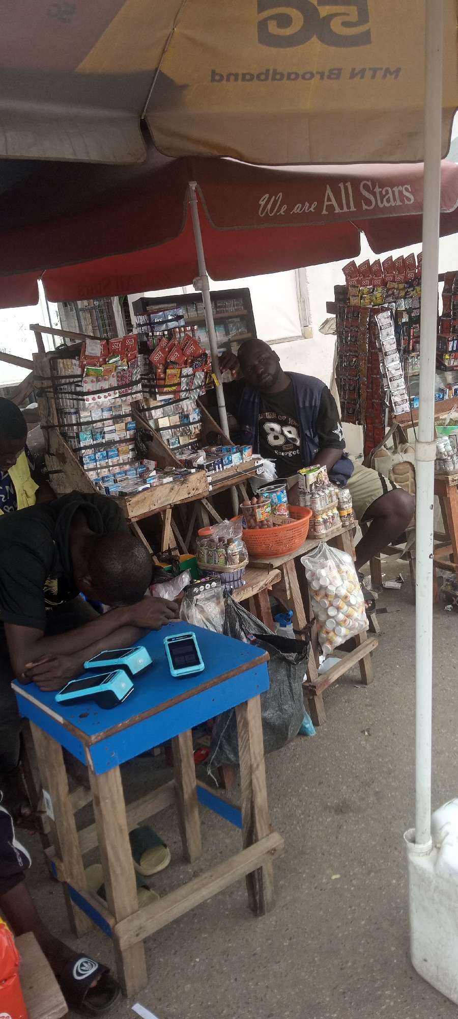 The image depicts an outdoor stall likely located in Jalingo, Nigeria, characterized by the presence of various goods, primarily cigarette packs, displayed on wooden shelves. A large umbrella, possibly a branded "MTN Broadband" and "We are All Stars" canopy, provides shade. Two men are visible; one appears to be sleeping or resting with his head on his arms on a blue table which holds three electronic devices, possibly Point of Sale (POS) terminals. The second man is seated, leaning back against the stall, looking towards the camera. The stall is laden with numerous packs of cigarettes arranged in stacks and hanging displays. Other items, such as small containers and a basket of what appear to be snacks or food items, are also present. The ground is paved, and a white plastic container is visible on the right side of the frame. The overall impression is of a busy street-side vendor stall, possibly in a marketplace.