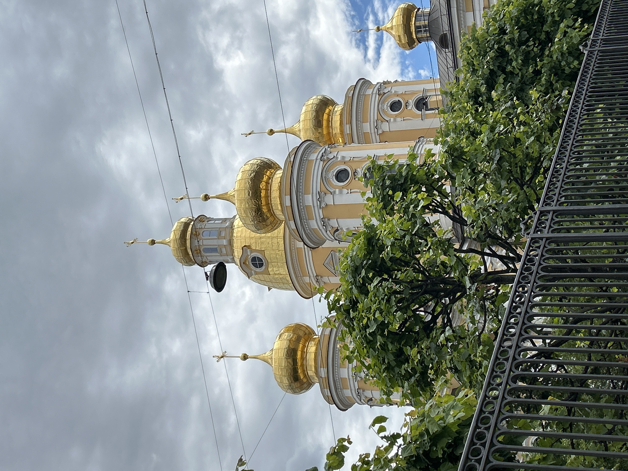 The image depicts a low-angle, slightly tilted view of the golden domes and spires of a church, likely a cathedral, against a cloudy sky. The architecture is ornate, with pale yellow walls and white detailing. Multiple domes, some larger and rounded, others more elongated and conical, are visible, each topped with a cross. The sunlight catches the gold, giving the structures a radiant appearance.
A dense, lush green tree partially obscures the lower portion of the church and also runs along the right side of the frame. A dark, wrought-iron fence with an intricate, repeating pattern runs along the far right, further framing the scene. The fence suggests the viewpoint is from a public space or walkway.
The sky is overcast with dramatic, grey clouds, interspersed with patches of brighter, blue sky. This indicates the weather is mixed, possibly threatening rain, but with breaks of sunshine. The lighting suggests it is daytime, though the exact time is hard to determine due to the cloud cover. There are no people visible in the image. The overall impression is one of grandeur and historical architecture, softened by the presence of nature. No visible text is present in the image.