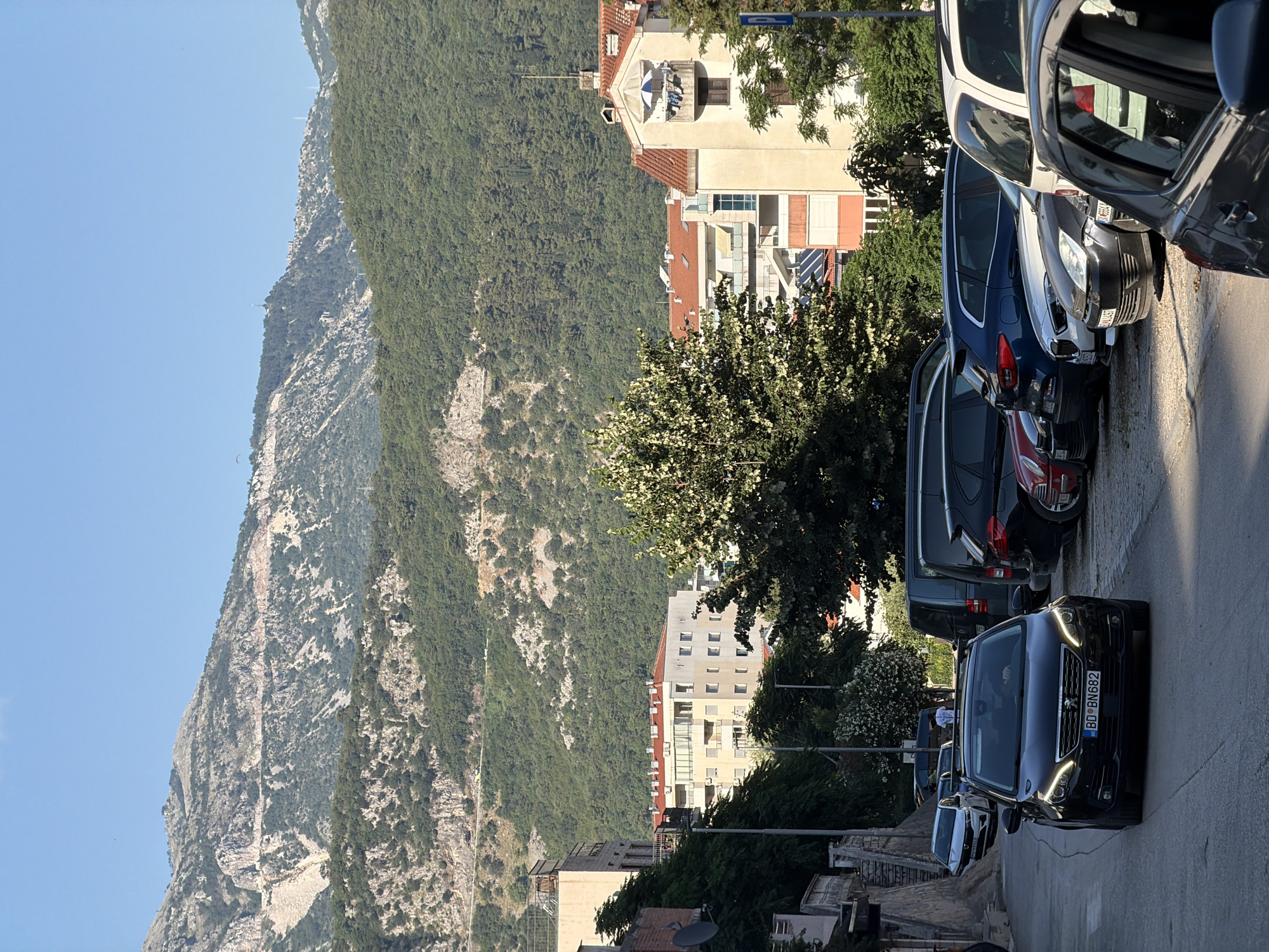 This is a vertical, medium shot photograph of a street in Budva, Montenegro, on a clear, sunny day. In the foreground, a black Peugeot 308 is driving down a paved street, with its headlights on. The car has a license plate that reads "BD BN682". To the right of the street, several cars are parked along the curb, including a white SUV, a blue station wagon, a silver sedan, and a red car. Behind the parked cars, there is a cluster of buildings with red-tiled roofs. In the background, lush green hills rise up, with rocky outcrops and sparse vegetation. The sky is a clear blue. There are no people clearly visible in the image beyond the driver of the Peugeot.