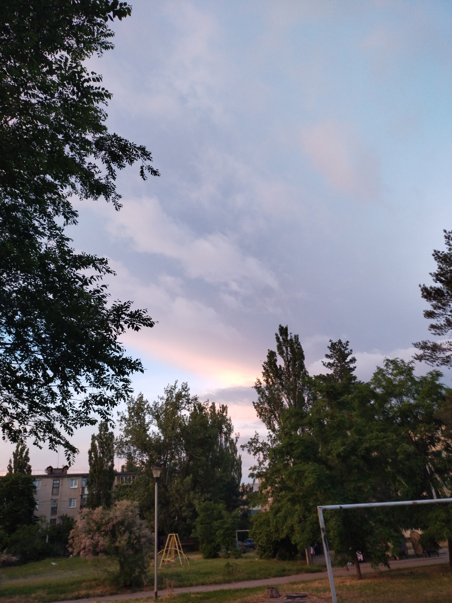 The image depicts an outdoor scene in Horishni Plavni, Ukraine, at what appears to be dusk. The sky is a pale blue with streaks of pink and white clouds, suggesting the sun has recently set or is about to set. In the foreground, there is a grassy area with some dry patches, and a dirt path runs horizontally across the bottom. To the left, the dense foliage of a tree with dark green leaves partially obscures the view of a multi-story apartment building. The building is made of light-colored brick and has several windows. To the right of the building, a cluster of tall, slender trees with dark green foliage stands out. Further to the right, a street lamp with a gray pole is visible. A small, yellow swing set structure is situated in the middle ground. In the background, more trees and the tops of other buildings are visible. The overall impression is a residential area at the end of the day.