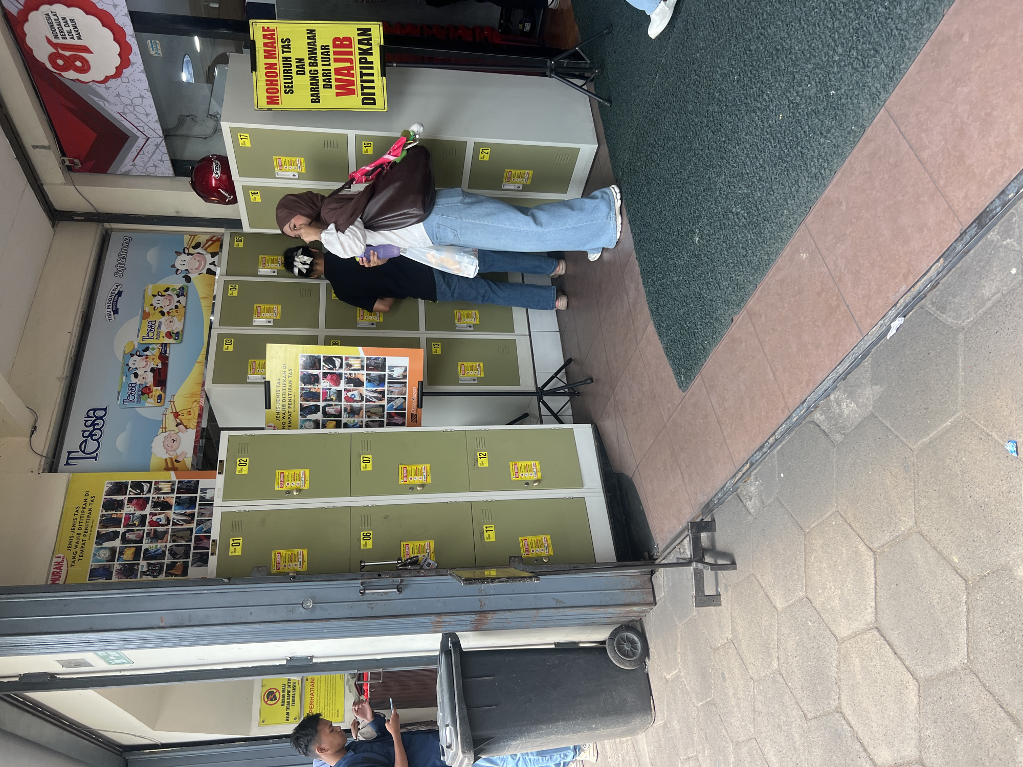 The image depicts a street-level view of a store entrance in Yogyakarta, Indonesia, characterized by a row of green lockers, presumably for customer use, and several large posters. In the foreground, a dark grey trash bin with wheels stands near a metal gate. To the left, a young man wearing a blue shirt and jeans is looking at his phone, and to the right, two women are standing near the lockers, one of whom is wearing a brown hijab and a large brown bag. The lockers are numbered sequentially from 01 to 21 and appear to be part of a self-service system. Behind the lockers, various posters are displayed, including one with images of different types of bags and another advertising "Tessa" tissues. A red motorcycle helmet is visible on top of one of the lockers. The ground is paved with hexagonal tiles, and a green mat is placed at the entrance. The overall setting suggests a busy commercial area where customers might need to store their belongings before entering the store.