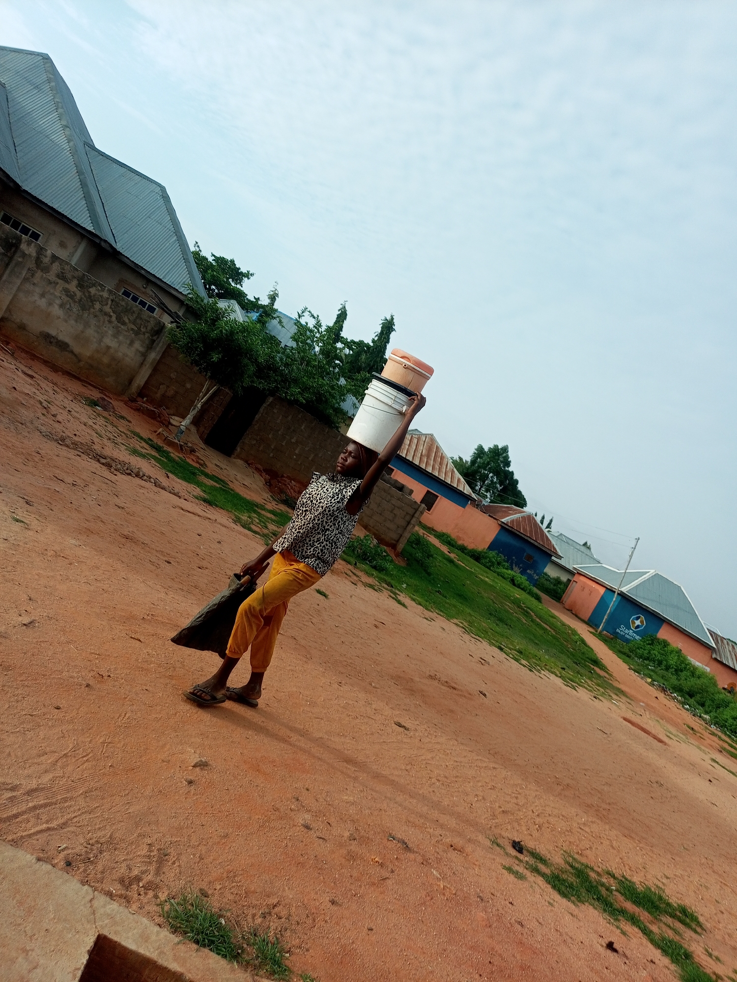 In Tumfure, Nigeria, a young person is depicted carrying two buckets stacked on their head and a dark bag in their right arm. They are wearing a patterned short-sleeved shirt and yellow pants, with sandals on their feet. The scene unfolds on a dirt path, with a row of buildings in the background. The architecture of the buildings includes corrugated metal roofs and some feature colorful painted facades, including one with a "StarTimes" logo. The sky is overcast.
