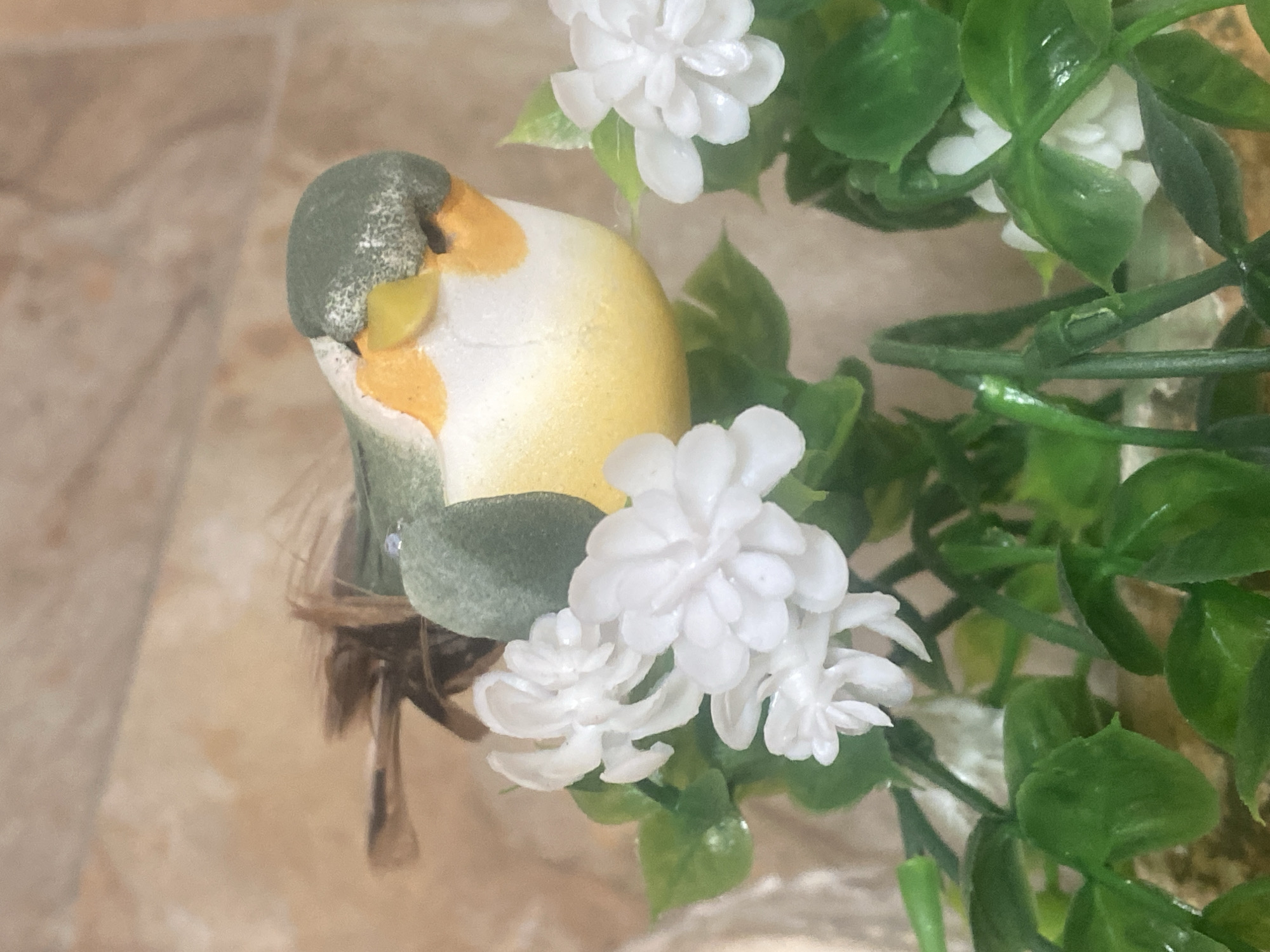 The image presents a close-up view of a decorative arrangement, likely an indoor setting in Novokuznetsk, Russia. The central subject is a small, artificial bird figurine, characterized by a grey-green head, a prominent yellow patch around its eye and beak area, and a predominantly white and yellow body. The figurine appears to have a textured, somewhat fuzzy surface.
The bird is nestled within a small, fibrous structure resembling a nest, which includes hints of brown feathery material. Surrounding the figurine and nest is artificial foliage featuring clusters of small, multi-petaled white flowers, possibly designed to mimic jasmine, alongside glossy green leaves. The plastic stems of the artificial plant are also visible.
The background consists of a light brown or beige tiled surface, which is slightly out of focus, suggesting it could be a floor or a table. The lighting is soft and even, indicating an interior setting. There are no human subjects or activities taking place. No discernible text is visible within the frame. The image captures a decorative still life.