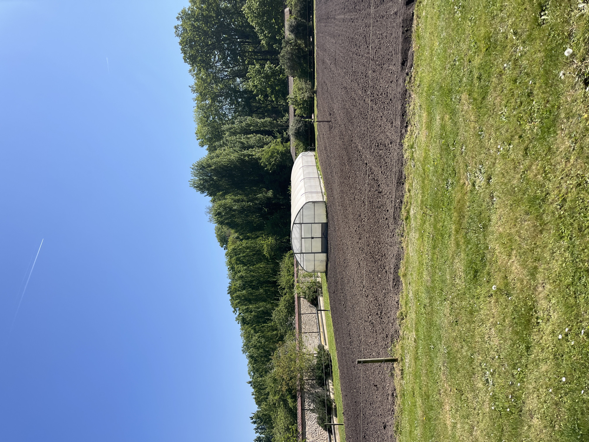 The image shows a rural scene in Saint-Rémy-lès-Chevreuse, France. On one side is a freshly tilled field, bordered by lush green grass on the right. A white, arched greenhouse stands adjacent to the field. In the background is a dense line of trees. A partially visible stone wall with fencing runs parallel to the field. Above, a clear blue sky is visible.