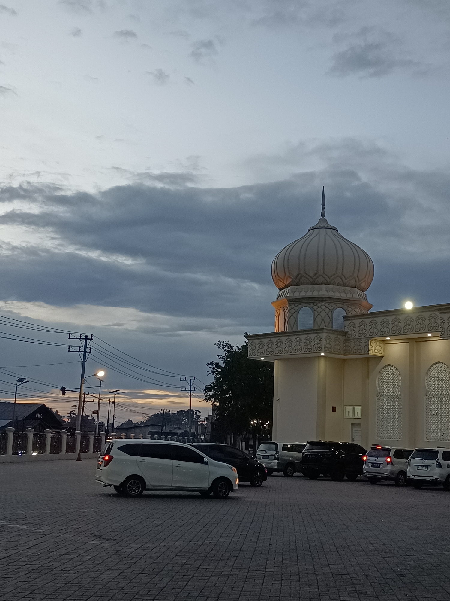 The image captures a mosque in Mee Tanjong Usi, Indonesia, during dusk. The sky is filled with dramatic, layered clouds in shades of gray and white, with a hint of warm light breaking through near the horizon on the left. The mosque's prominent dome, topped with a spire, is a light tan color and is illuminated by artificial lighting, casting a warm glow. The mosque building itself is cream-colored with decorative patterned windows and intricate detailing along the roofline. Several vehicles, including white vans and a black SUV, are parked in the paved courtyard in front of the mosque. Power lines and streetlights are visible in the background to the left, indicating the presence of infrastructure within the town. The ground is paved with dark gray cobblestones. The overall scene is serene, with the fading light and the imposing structure of the mosque creating a sense of quiet reverence.