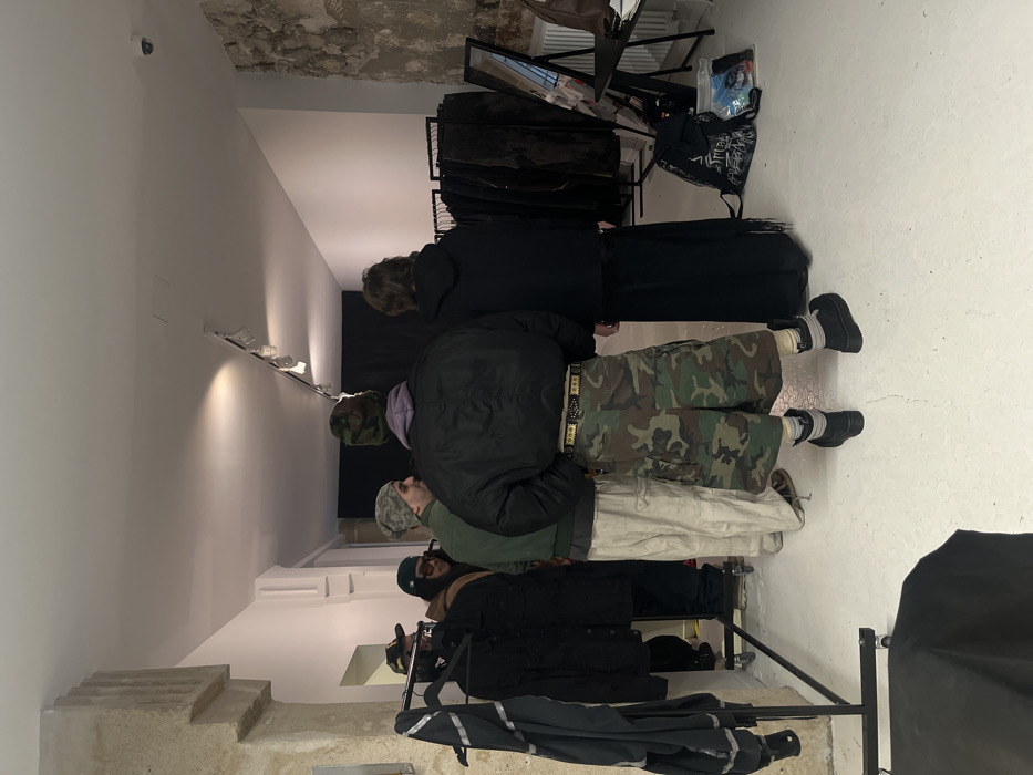 In an interior space with exposed brick walls and a smooth, white floor, a group of people stand observing clothing racks filled with various garments. One person is wearing camouflage pants and a black jacket, while another wears white pants with a similar jacket. A person in an all-black outfit stands near the back. The room is lit by track lighting and natural light, casting shadows and highlighting the textures of the clothes.