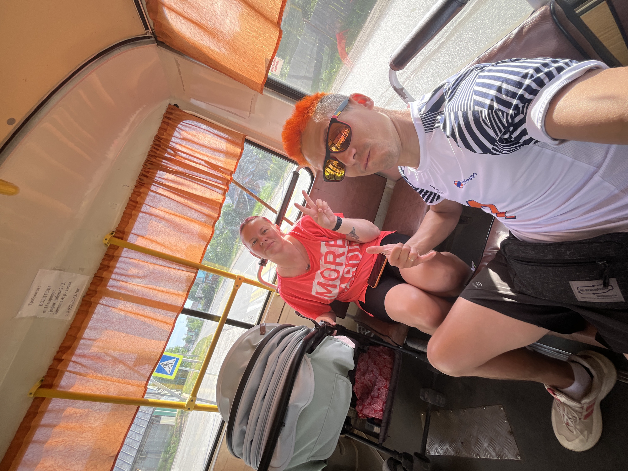 Here is a forensic description of the image:
Two individuals, likely tourists, are inside what appears to be a bus or trolleybus. The man has orange hair and sunglasses, wearing a white athletic shirt. The woman is wearing a coral-colored T-shirt, black shorts, and has a tattoo on her arm. They are both making hand gestures, appearing to be posing for a selfie. A baby stroller is visible next to the woman.
The bus interior has orange curtains. Through the window, a street scene is visible, including a pedestrian crossing sign. A recruitment poster for bus drivers is visible inside the bus. The text from the poster is: "Требуются: ВОДИТЕЛИ на 15 маршрут. График работы 2/2, Зарплата 4т.р.- смена. τ. 8-922-36-63-108" (Translation: "Required: DRIVERS for route 15. Work schedule 2/2, Salary 4t.r. - shift. T. 8-922-36-63-108"). Another poster with the text "ОКНО РАЗБИТЬ Молотком" (Translation: BREAK WINDOW Hammer") is also visible.
Based on the Russian text, the location is likely in a Russian-speaking area, possibly the city of Chaikovsky, Russia, although this cannot be confirmed.