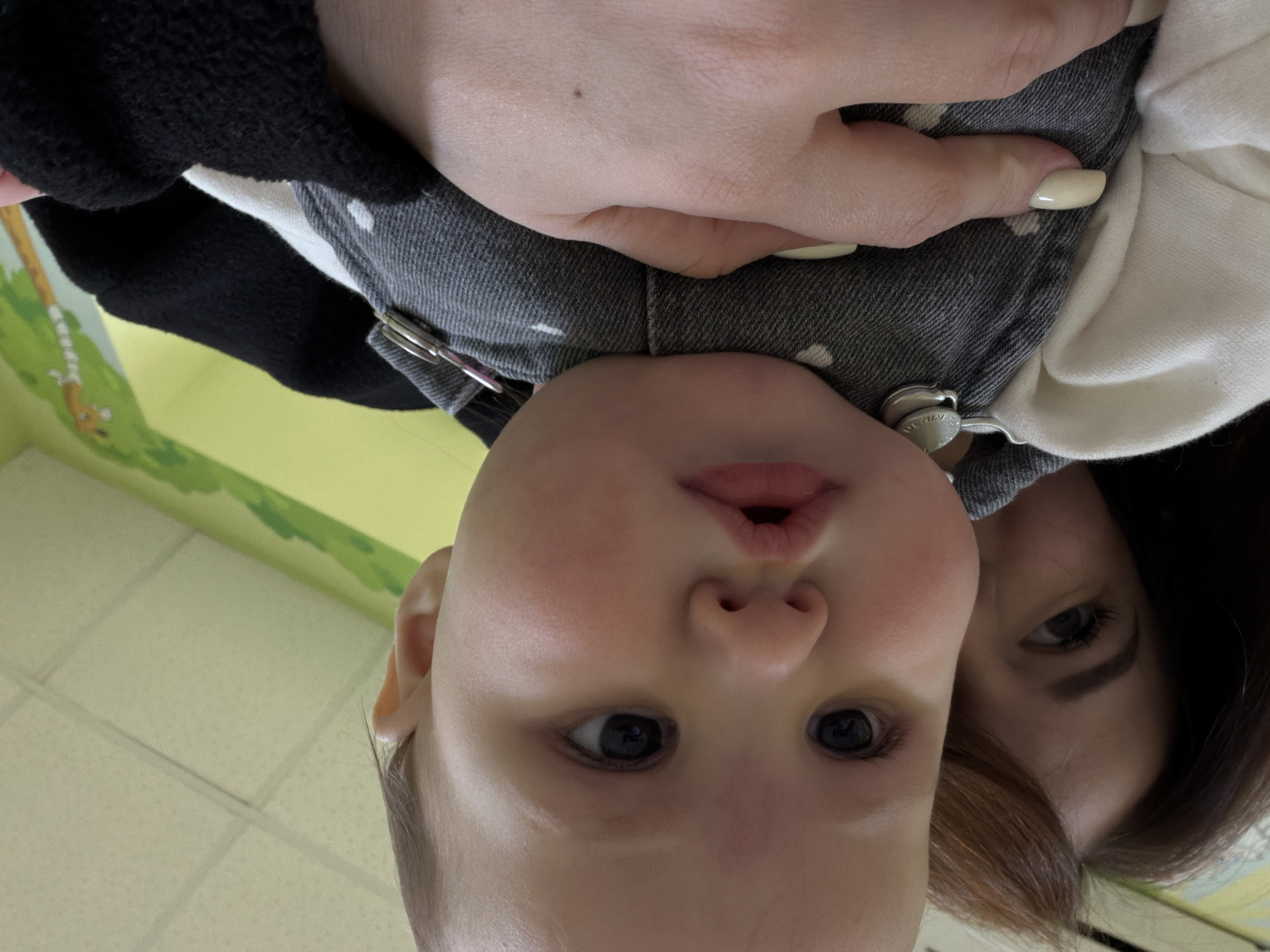 A close-up, slightly angled image features a baby being held. The baby has fair skin, dark eyes, and a slightly puckered mouth. The baby is wearing a gray and white garment with metal fasteners. A woman is partially visible behind the baby; only her eye, eyebrow, and some of her brown hair are in the frame. A hand with light-colored fingernails is also present. The background includes a section of wall with a green and yellow design, and a tiled ceiling. Location information is unavailable.