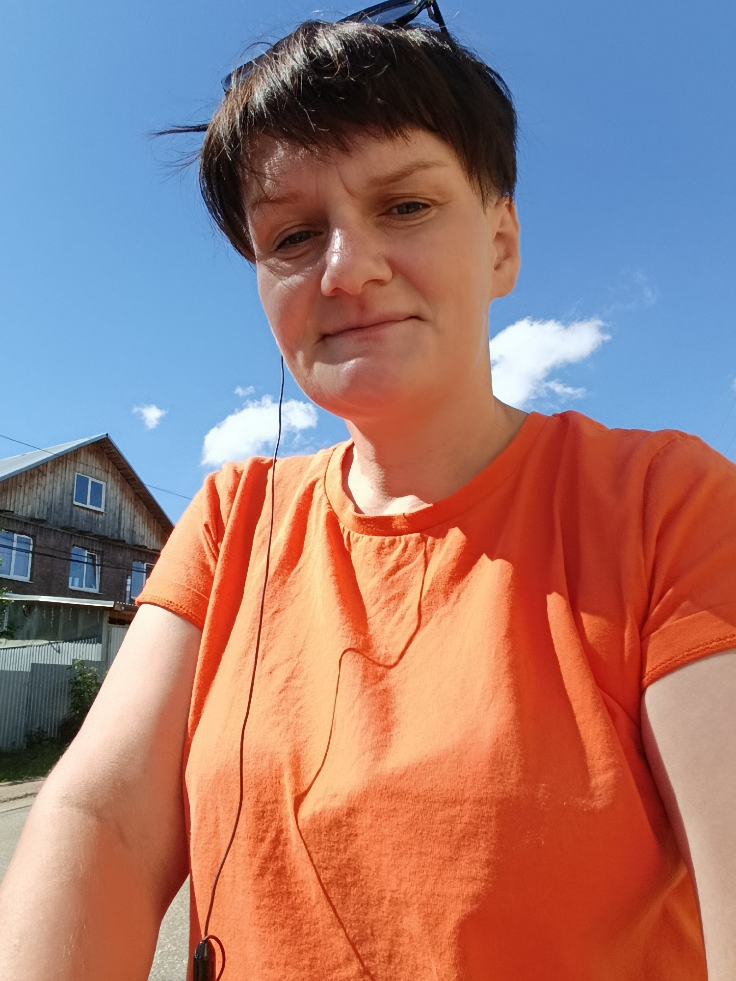 A woman is taking a selfie outdoors. She has short, dark hair and is wearing an orange t-shirt and sunglasses on her head. She is also wearing earbuds. In the background, there is a wooden house with windows and a blue sky with some clouds. The location appears to be in the city of Foky, Russia, as indicated by the style of the house and the general environment.