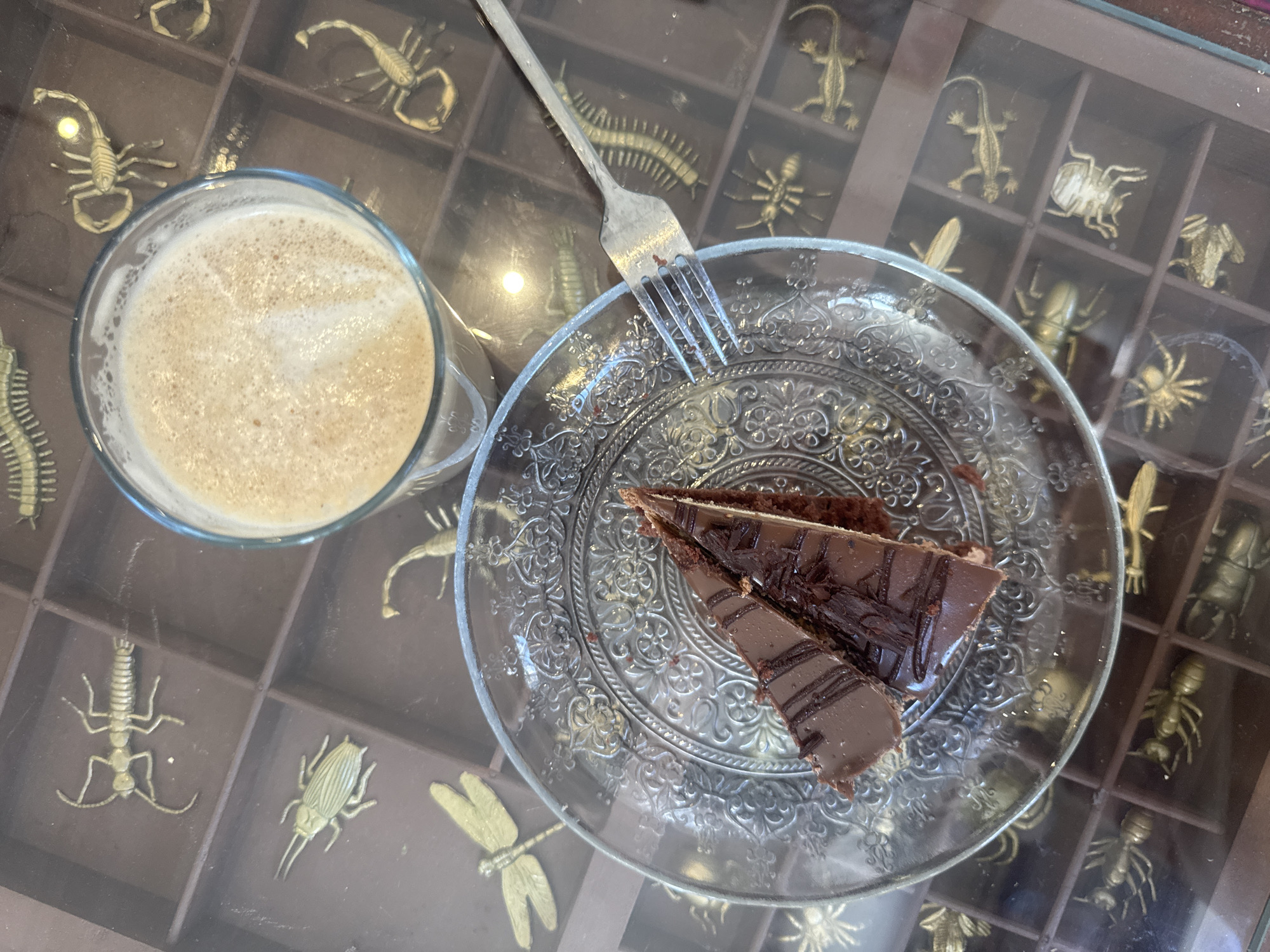 A high-angle shot shows a coffee drink in a clear glass mug and a slice of chocolate cake on a clear, decorative plate with a fork on a tabletop, likely inside a cafe. The tabletop is unique; it is a divided tray showcasing numerous small, golden-colored insect and arachnid figures (e.g., scorpions, spiders, beetles). While the city and specific location are not visually discernible from this image alone, the presence of this image suggests it was taken in Tel-Aviv, Israel.