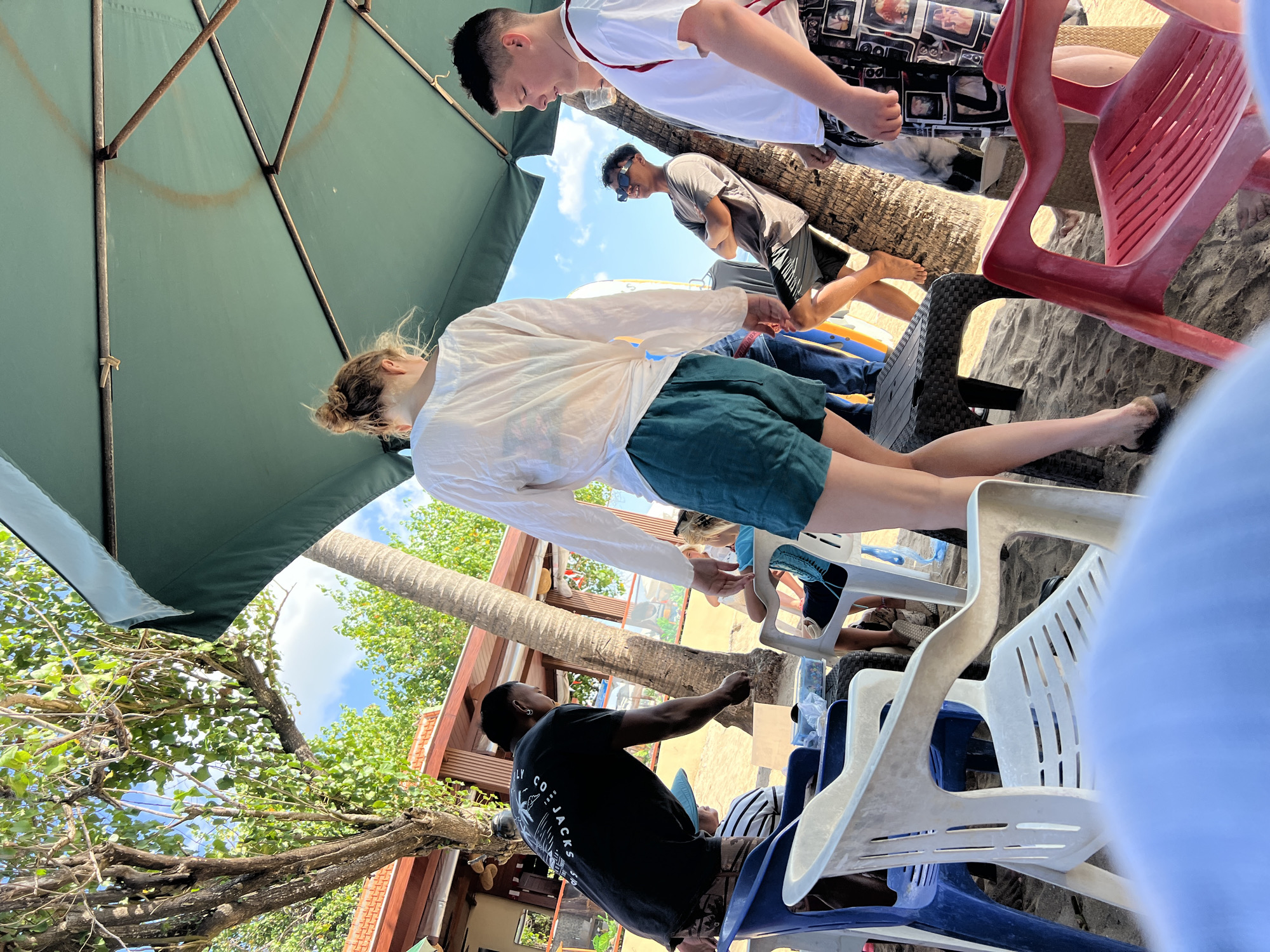 In Kuta, Indonesia, under a large green umbrella providing shade on a sandy beach, a group of people are gathered. A young woman with her hair in a bun, wearing a white long-sleeved shirt and teal shorts, stands facing away from the camera. To her left, a man in a black t-shirt with a graphic print, possibly "CO-JACKS 1998," is leaning forward, looking down at something. He has an earring and is wearing patterned shorts. To the right of the woman, a young man with sunglasses, wearing a grey t-shirt and dark shorts, leans against a palm tree. Further to the right, another young person in a white t-shirt and patterned shorts is visible. Several plastic chairs, some blue and white, and a red chair, are scattered on the sand. In the background, a building with a reddish-brown roof and palm trees can be seen, suggesting a beachside establishment. The sky is blue with some clouds. The scene appears to be a casual gathering on a sunny day.