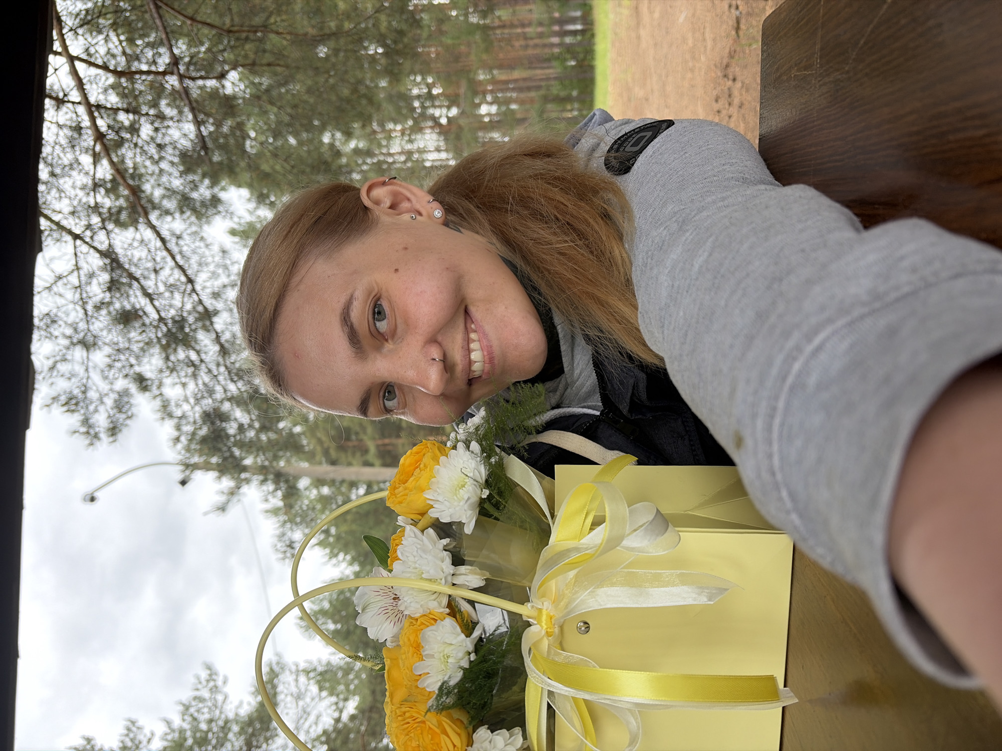 A woman with fair skin and light brown hair pulled back in a ponytail is posing for a selfie with a bouquet of yellow and white flowers in a yellow gift bag. She is wearing a gray hooded sweatshirt. The photo is taken outdoors, possibly at a picnic table, with trees visible in the background. A street lamp is also visible in the background. The location appears to be Chaykovsky, Russia.