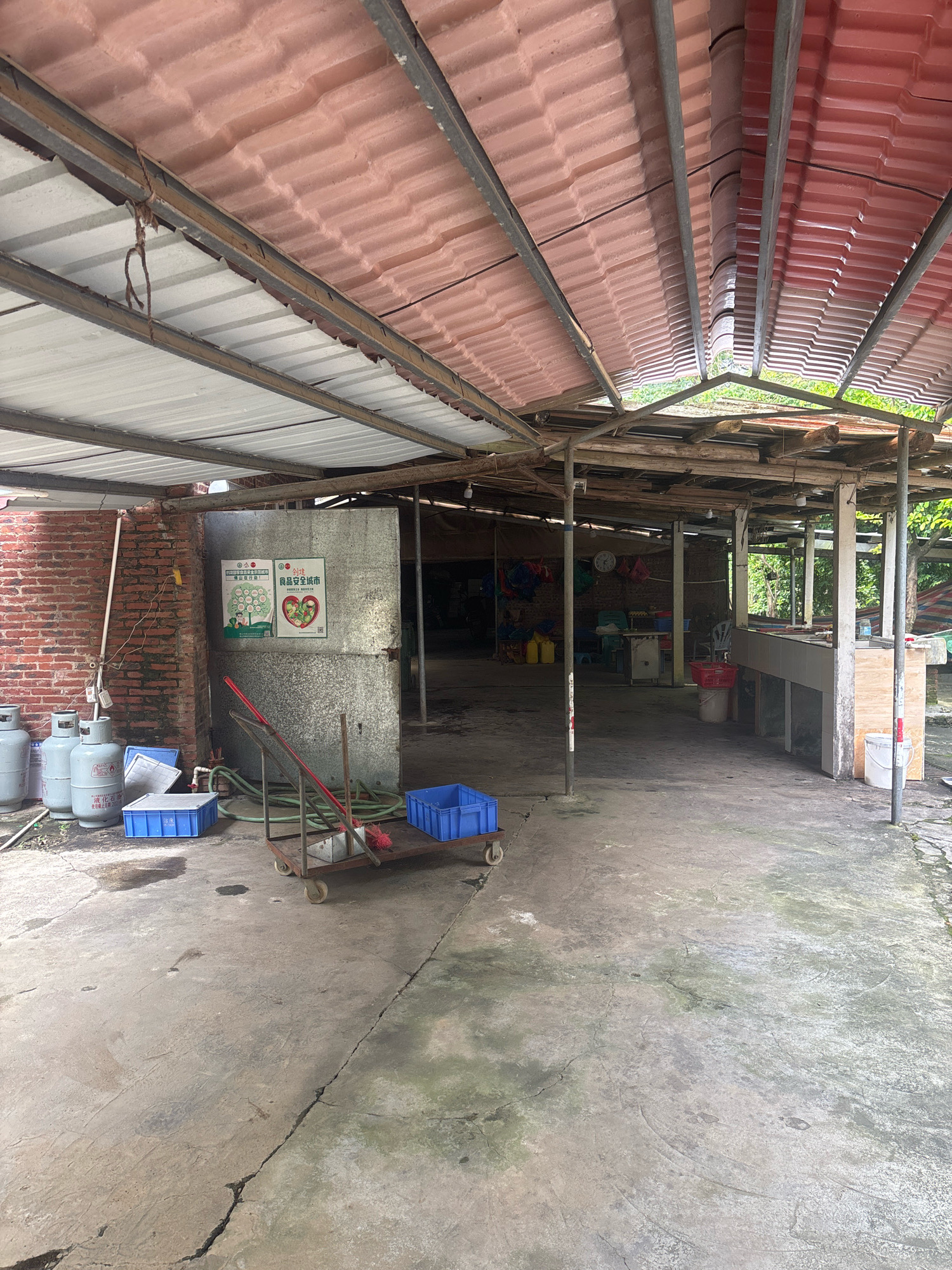The image depicts an outdoor storage or work area, likely part of a market or processing facility, located in the city of Sanshui District, China. The scene is covered by a corrugated metal roof supported by metal and wooden beams. A concrete floor, showing signs of wear and some moss growth, dominates the foreground and midground.
On the left side of the image, several large propane gas tanks are visible. Adjacent to them are blue plastic crates and a handcart holding a broom and a red object, possibly another cleaning tool. A brick wall forms the backdrop on the left. Two posters are affixed to a galvanized metal surface, one featuring a circular diagram with illustrations of healthy fruits and vegetables, and the other displaying a heart shape composed of fruits with text in Chinese.
The background reveals an open-sided structure with various items stored within, including bags of yellow liquid, hanging bags, a clock on a wall, and what appears to be a workbench or counter area with sinks. Greenery is visible beyond the structure. The overall impression is of a functional, utilitarian space that is not meticulously maintained.