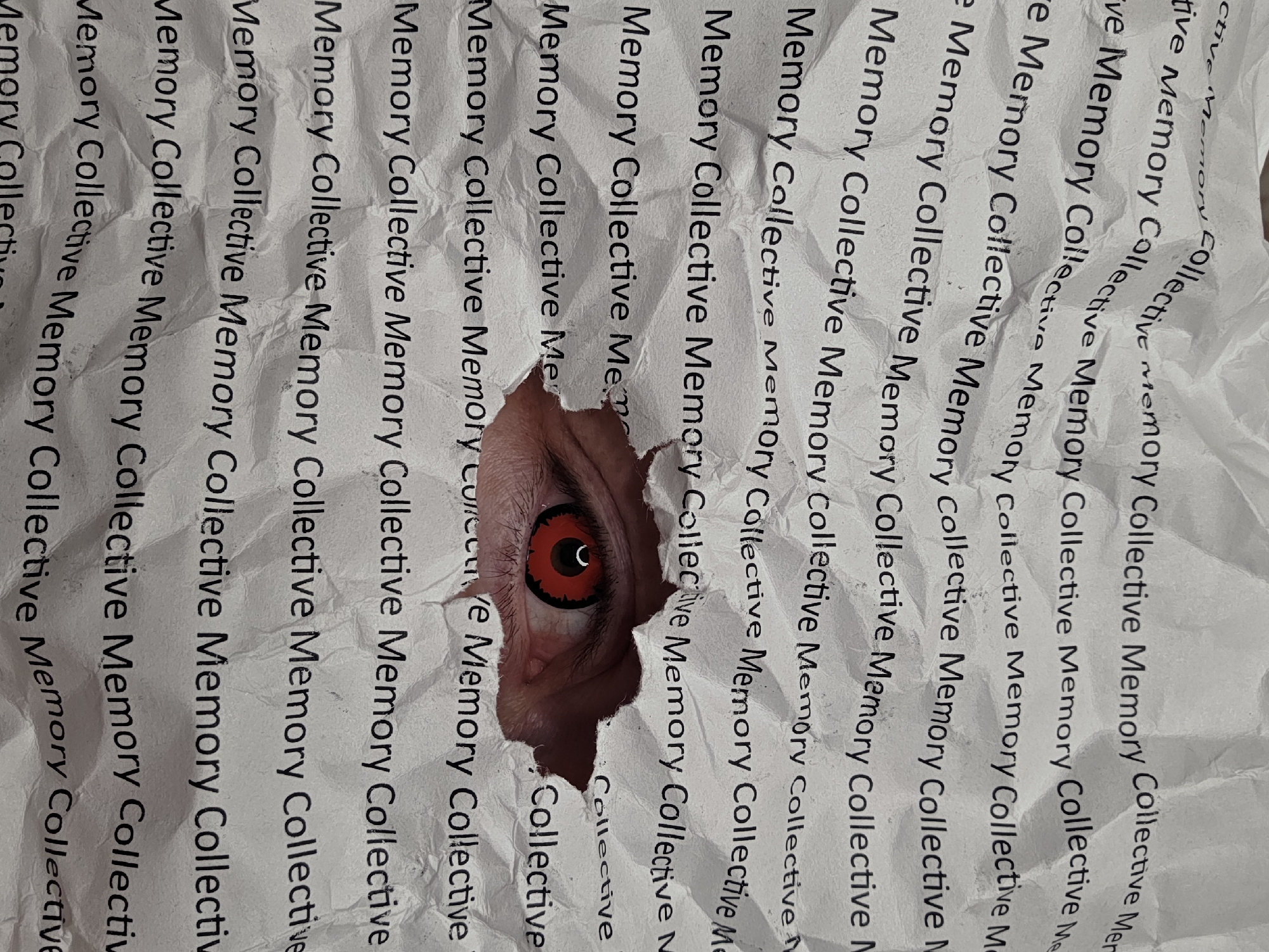 A sheet of paper, printed repeatedly with the phrase "Memory Collective Memory Collective," is crumpled and torn to reveal a human eye with a red sclera underneath. The location of the image, Chaikovsky, Russia, is not revealed within the image itself.