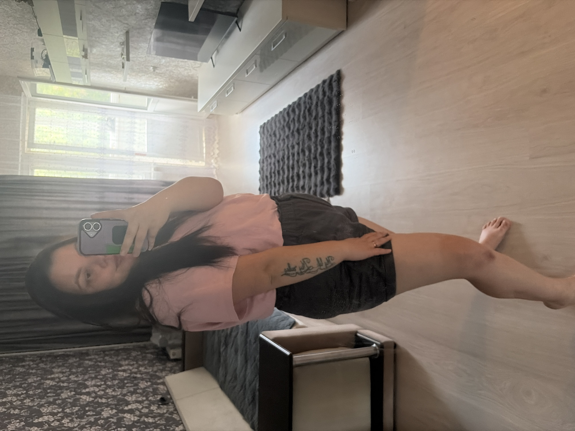A woman is standing in a room, taking a mirror selfie with her phone. She is wearing a pink t-shirt and dark shorts. She has a tattoo on her left arm. The room appears to be a bedroom or living area, with a bed or sofa on the left, a rug on the floor, and a TV stand with a television on the right. There is a window and a balcony door in the background, letting in natural light. The flooring is light-colored wood. The image was taken in Izhevsk, Russia.