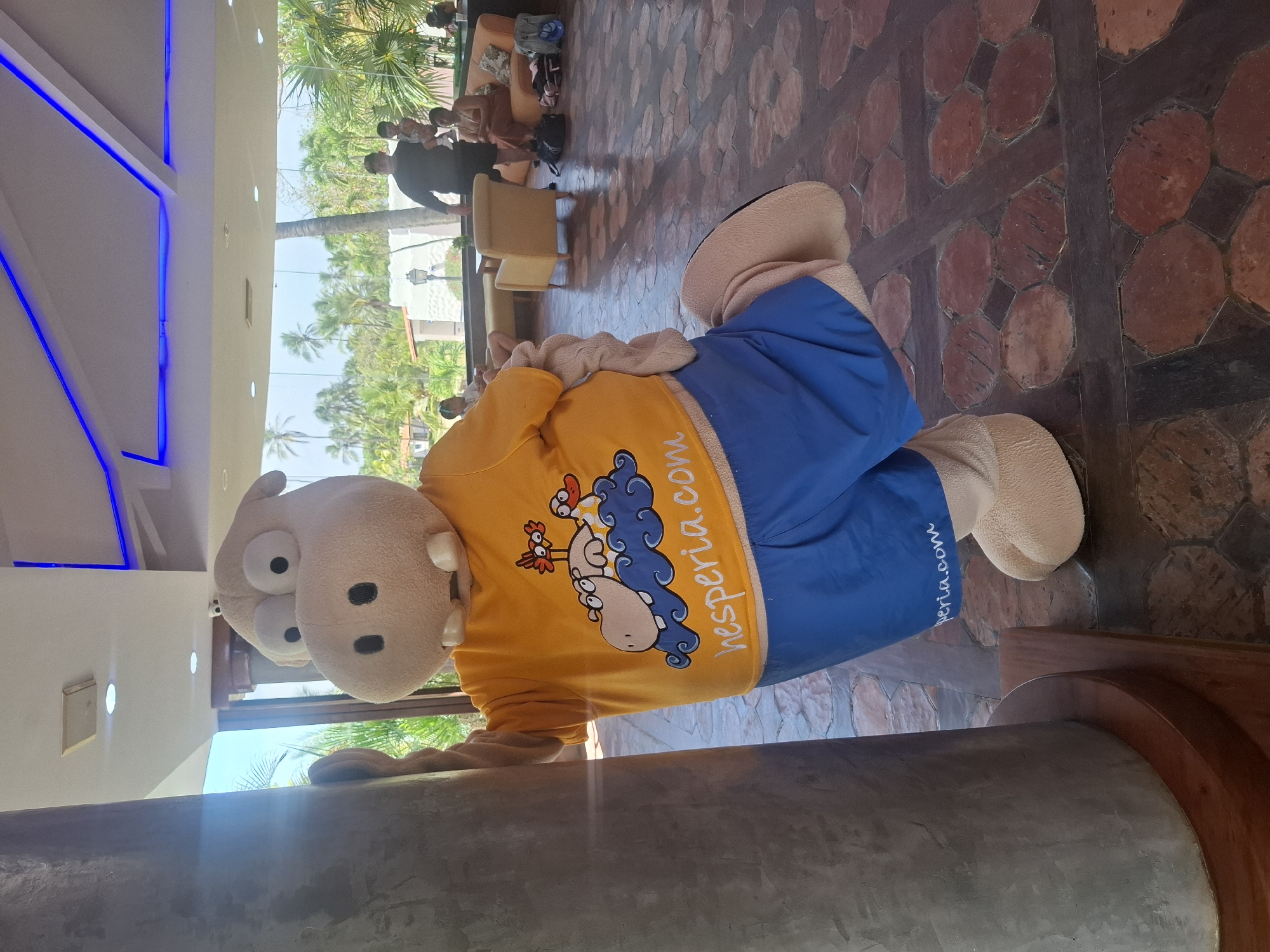 A large mascot costume, appearing to be a light brown hippopotamus with big eyes and buck teeth, stands in what seems to be a hotel lobby or common area in Playa El Agua, Venezuela. The mascot is wearing a bright yellow t-shirt with a cartoon image of two birds and a hippopotamus on it, along with blue shorts. The t-shirt has the text "hesperia.com" written on it in white. The mascot is leaning against a large, gray pillar, with its right leg bent at the knee and extended backward. In the background, through large windows, palm trees and lush greenery are visible, suggesting a tropical location. Inside the lobby, there are people sitting on furniture, including a man holding a child. The floor is tiled with reddish-brown octagonal tiles. The lighting suggests it is daytime, with sunlight streaming in and artificial lights on the ceiling. The overall scene appears to be a public area within a resort or hotel, likely at the Hesperia resort given the text on the mascot's shirt.