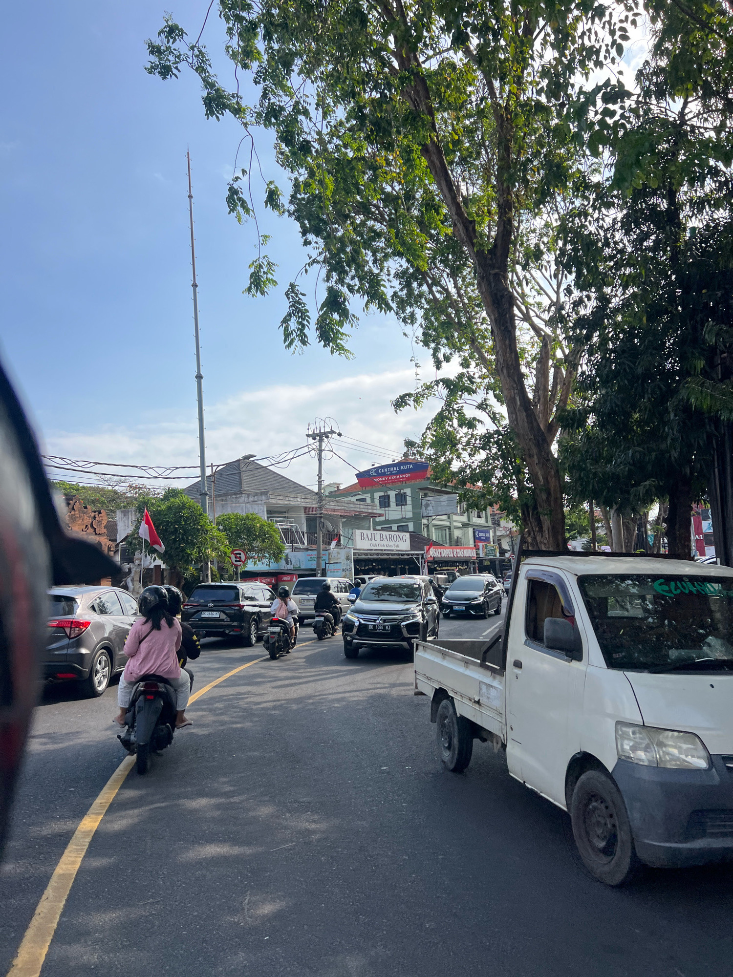 This is a street scene in Kuta, Indonesia, on a clear, sunny day. The image captures a busy road with various vehicles and pedestrians.
In the foreground, the left side of the frame shows a blurred view of a person's helmet, suggesting the image was taken from the perspective of someone riding a motorcycle. To the right, a white pickup truck is prominently positioned, partially obscuring the view of the road ahead. The truck has a dirty exterior and visible wear and tear.
The road is asphalt and marked with a yellow line. Several vehicles are on the road, including cars and motorcycles. A grey SUV is visible on the left, and a black SUV is in the middle lane. Two people on a motorcycle are in the left lane, with the passenger wearing a pink jacket and white pants. Another motorcycle with two riders is further ahead in the same lane.
In the background, buildings line the street. One building has a sign that reads "CENTRAL KUTA MONEY EXCHANGE." Another sign says "BAJU BARONG." A red and white Indonesian flag is displayed on a pole. Trees with green foliage are present along the sides of the street and overhead. A tall, thin pole, possibly a communication tower, stands against the blue sky. Overhead power lines are also visible. The sky is mostly clear with some scattered clouds. The overall impression is of a typical urban street scene in a tropical location, with traffic and commercial activity.