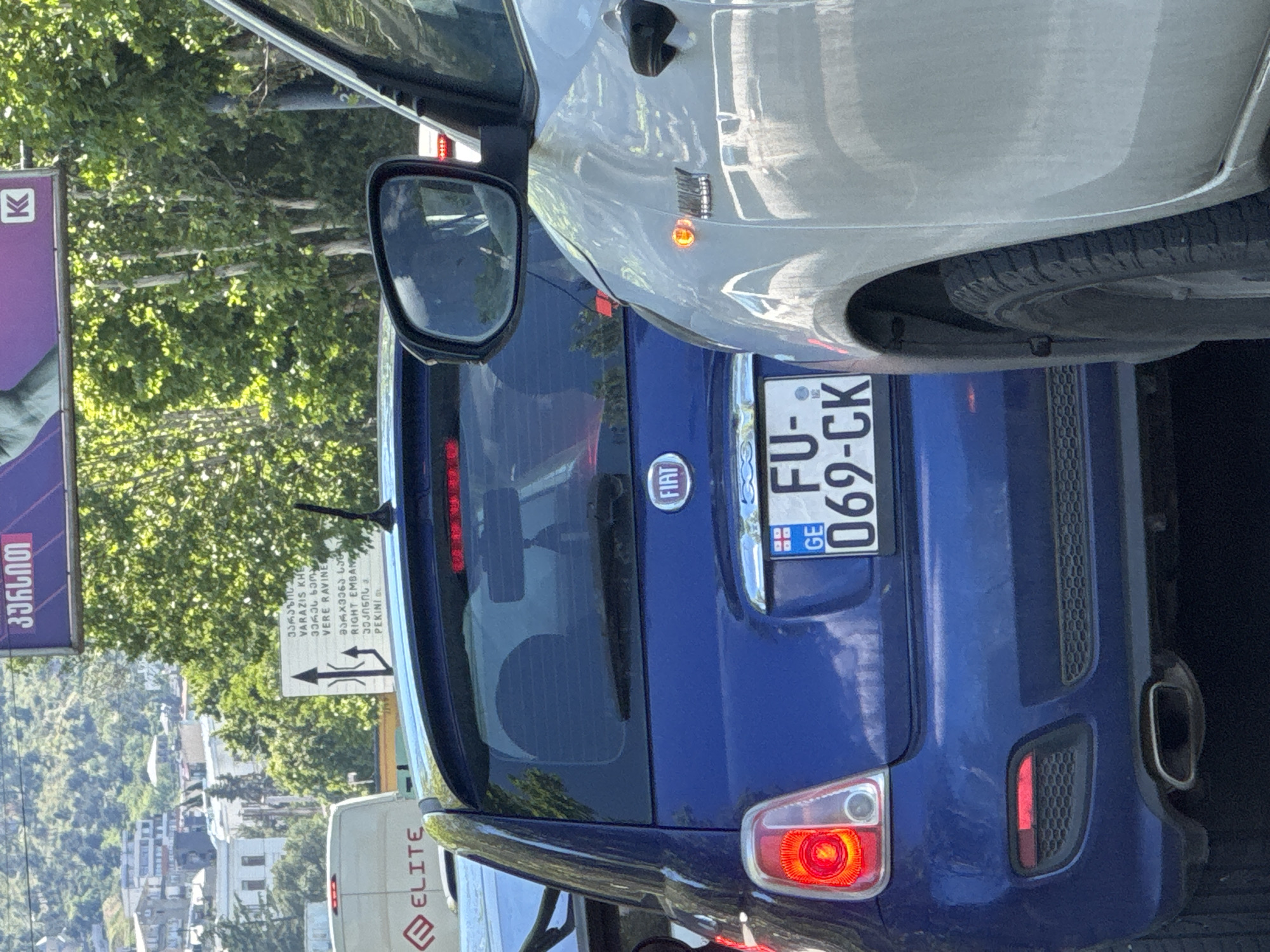 This media file captures a street scene, rotated 90 degrees clockwise, under bright daylight on a clear day. In the foreground, the rear of a silver or white car is visible, with its right side mirror and an illuminated amber turn signal on the front fender.
Behind the silver car is a blue Fiat 500, seen from the rear. Its license plate, "FU-069-CK", is clearly visible, featuring a "GE" country code and the Georgian flag emblem, indicating the location is Georgia. The Fiat's "FIAT" logo is centered on its tailgate, and its red taillights are visible. The exhaust pipe can be seen on the right side of the rear bumper.
To the left of the cars, a large purple billboard with Georgian text, including "კურსით" (kursit), and a prominent "K" logo is partially visible. Further back, a white traffic sign provides directional information in both Georgian and English. The English text reads "VARAZIS KHE (VERE RAVINE)", "RIGHT EMBAN", and "PEKINIG", confirming the Georgian location and suggesting it is likely Tbilisi. The sign also displays directional arrows, indicating options for going straight, turning right, or a merge/U-turn.
Lush green trees line the street, and in the distance, a city skyline with various buildings is visible. Part of a white van with the word "ELITE" printed on its side is also seen at the bottom left. The overall impression is a bustling urban environment.