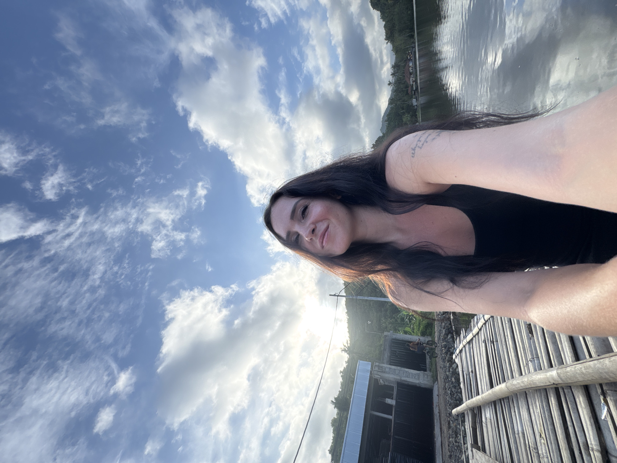The image captures a woman in a black tank top posing for a selfie from a bamboo structure on the water's edge. She has long, dark hair and a tattoo on her left arm. The background reveals a serene lake reflecting the sky, with lush green hills and a few buildings nestled among the trees. The sky is a canvas of blue with scattered clouds, suggesting a partly cloudy day. A building with a corrugated metal roof and a fence can be seen to the left of the frame, along with a person riding a bicycle in the distance. The overall scene evokes a tranquil, natural setting in Manggis, Indonesia.
