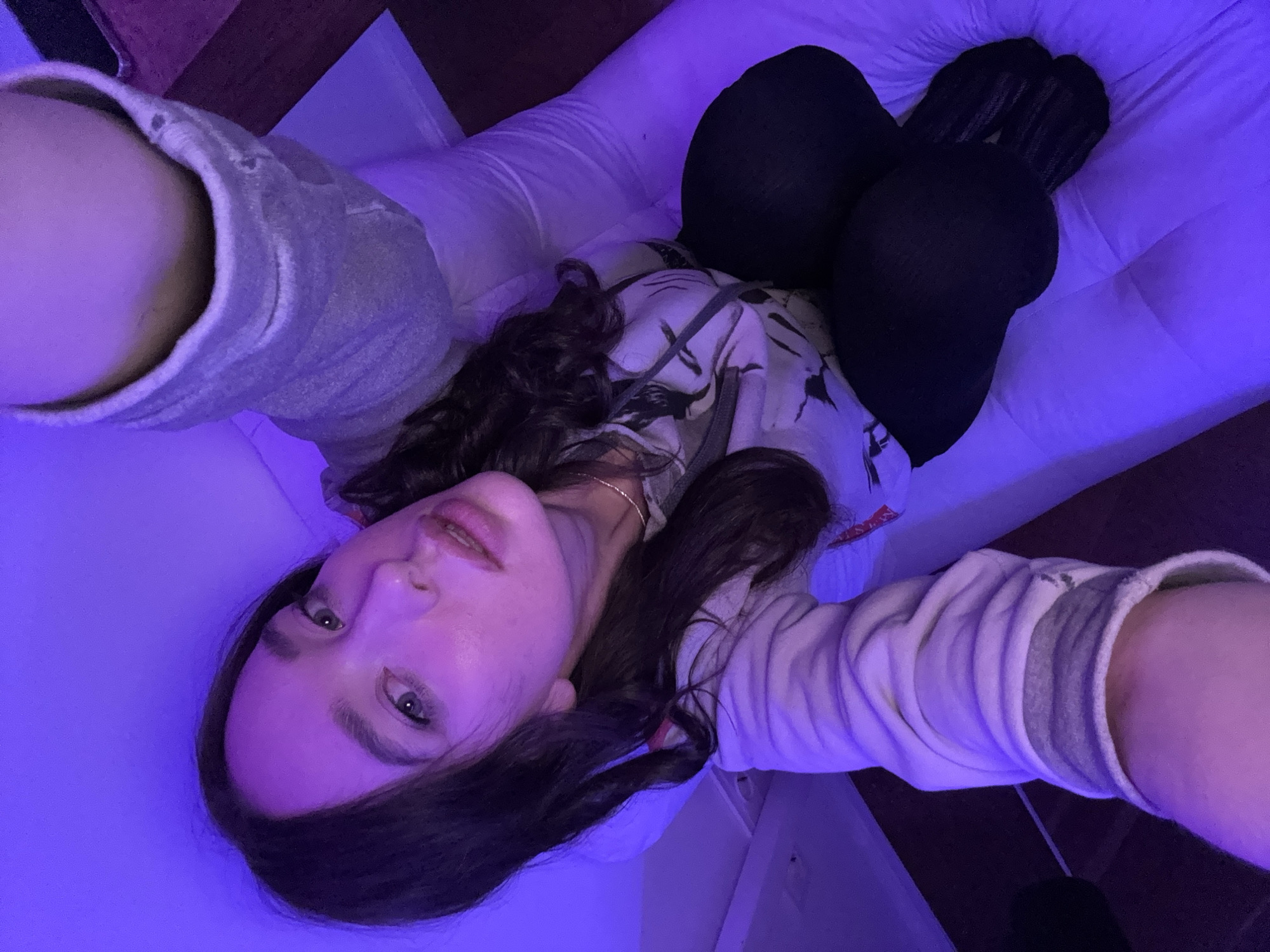 A low-angle selfie captures a young individual, likely female, lying down indoors. The scene is bathed in a distinct purple-blue ambient light, creating a moody and atmospheric setting, possibly from LED lighting.
The subject has long, dark, wavy hair cascading around her head and shoulders. She has light-colored eyes and full lips, looking directly at the camera with a neutral to slightly pensive expression. She is wearing a light-colored, possibly off-white or light grey, long-sleeved top or hoodie with a textured fabric. Beneath this, a patterned top with dark designs is visible. Her lower body is covered in dark leggings or pants, with dark socks visible on her feet. A thin, possibly gold-colored, necklace is subtly visible around her neck.
She appears to be lying on a bed or a couch, indicated by the white, quilted surface beneath her. The angle suggests she is holding the camera herself, with her arms extended towards her face. The background is simple, showing more of the white surface and some darker elements, possibly furniture or walls.
There is no visible text within the image. The time of day is not explicitly indicated but the artificial lighting suggests it could be evening or night. The provided context indicates the photo was taken in Moscow, Russia, though there are no visual cues within the image to confirm this location.