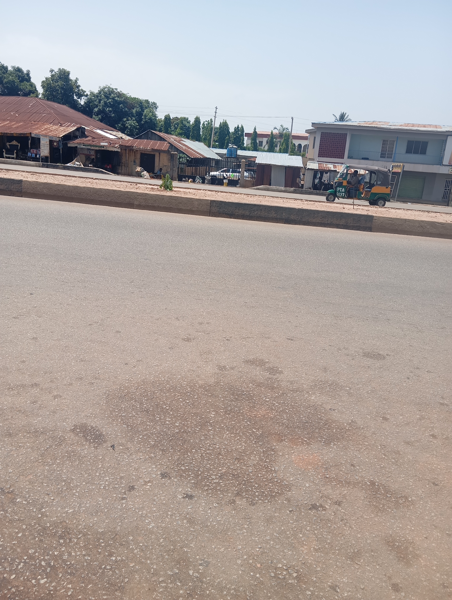The image captures a street scene in Jos, Nigeria, under a bright, clear sky, suggesting it is daytime and likely sunny. The foreground is dominated by a wide asphalt road, showing some wear and tear with scattered patches of discoloration. A concrete curb separates the road from a sidewalk area where a few structures and a three-wheeled vehicle are visible.
On the left side of the sidewalk, a cluster of buildings with corrugated metal roofs are visible. These appear to be shops or small businesses, with some displaying goods outside. One building has a sign that is partially obscured but appears to read "PRIME" followed by other text. Adjacent to these, more modern buildings are present, including one with a light blue facade and red patterned accents.
In the mid-ground, a green and yellow three-wheeled auto-rickshaw, commonly known as a "Keke," is driving on the street. It has passengers inside and displays text on its side which appears to be a license plate: "PTA 6497". Several other small, simple structures, possibly stalls or temporary shops, are situated along the sidewalk, some with people standing or sitting near them. A metal gate is also visible on the right side of this section.
The overall scene depicts a typical urban street environment, characterized by a mix of older and newer architecture, commercial activity, and local transportation. The bright lighting and clear sky indicate fair weather.