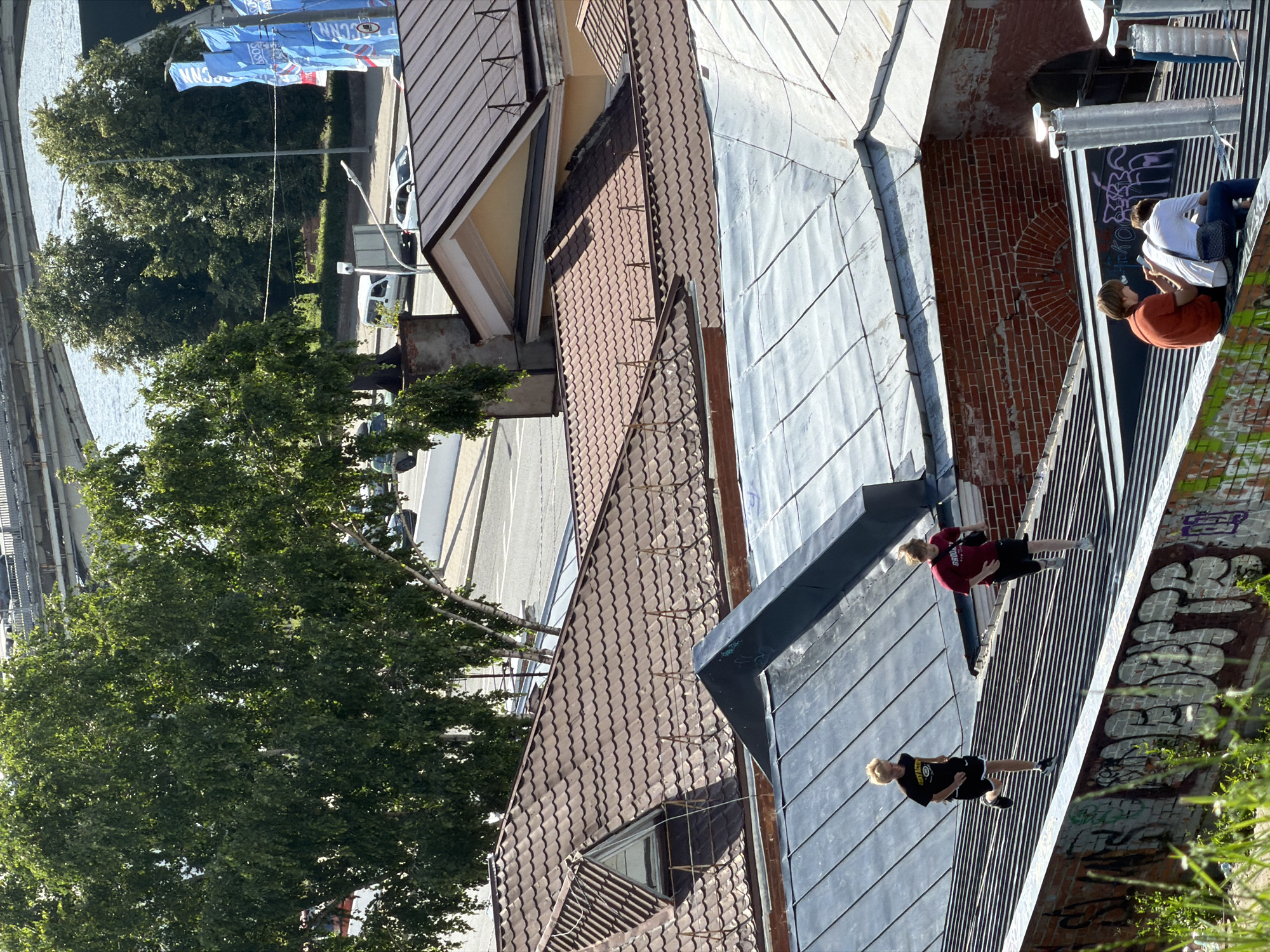 This aerial photograph, taken in Nizhny Novgorod, Russia, captures a cityscape dominated by rooftops and urban infrastructure. In the foreground, two young individuals, likely children or teenagers, are seen running on a dark, corrugated metal roof. One is wearing a black t-shirt with white lettering and black shorts, while the other wears a maroon t-shirt and black shorts, carrying a bag over their shoulder. They appear to be engaged in a playful activity or exercise.
Further in the background, several buildings with reddish-brown tiled roofs are visible, interspersed with sections of gray metal roofing. A prominent brick building with arched windows and some graffiti on its lower wall is situated between the two roofs. In the distance, lush green trees line a street where vehicles, including white vans and cars, are driving. A wide river flows behind the trees, crossed by a large bridge, suggesting a riverside urban environment. Blue banners with white lettering are displayed on poles near the riverbank. The scene is illuminated by bright sunlight, casting shadows across the rooftops. The overall impression is a dynamic urban setting with elements of both historical architecture and modern life.