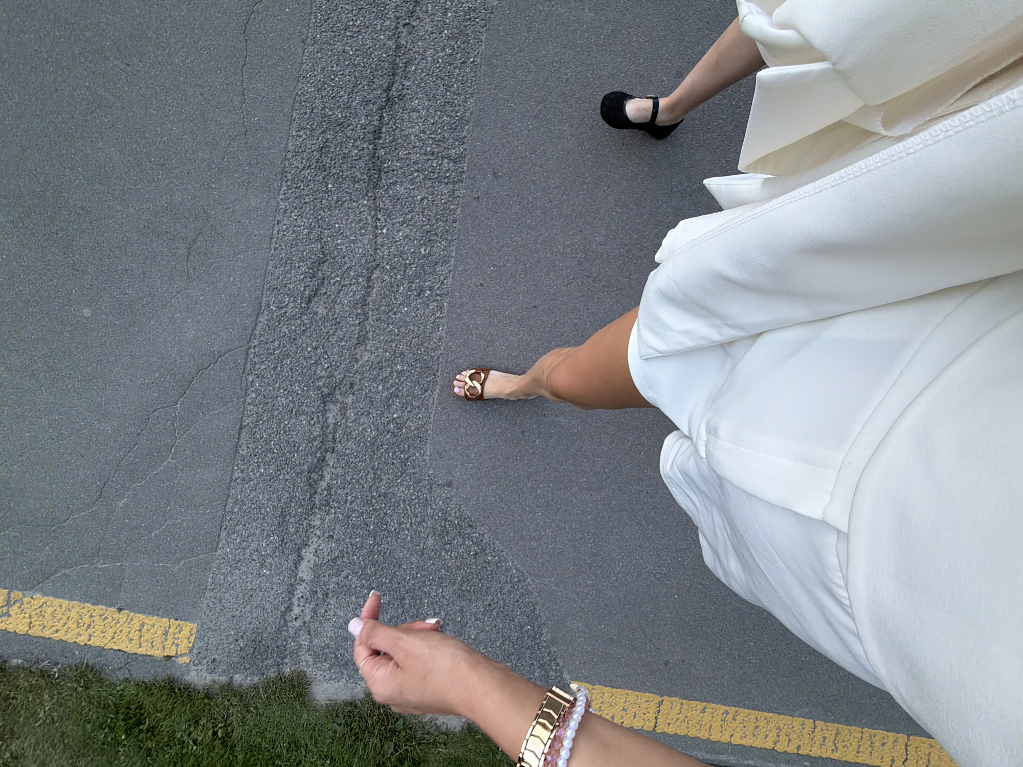 The media file shows a top-down perspective of two people walking on an asphalt path. One person is wearing black sandals and the other is wearing gold-chain sandals. Both individuals are wearing white clothing, appearing to be dresses or skirts. The path is bordered by a yellow line and grass. The lighting suggests it is daytime, likely outdoors. The scene is simple and focuses on the lower bodies and footwear of the individuals. There is no visible text. The location context is Polianytsia, Ukraine.